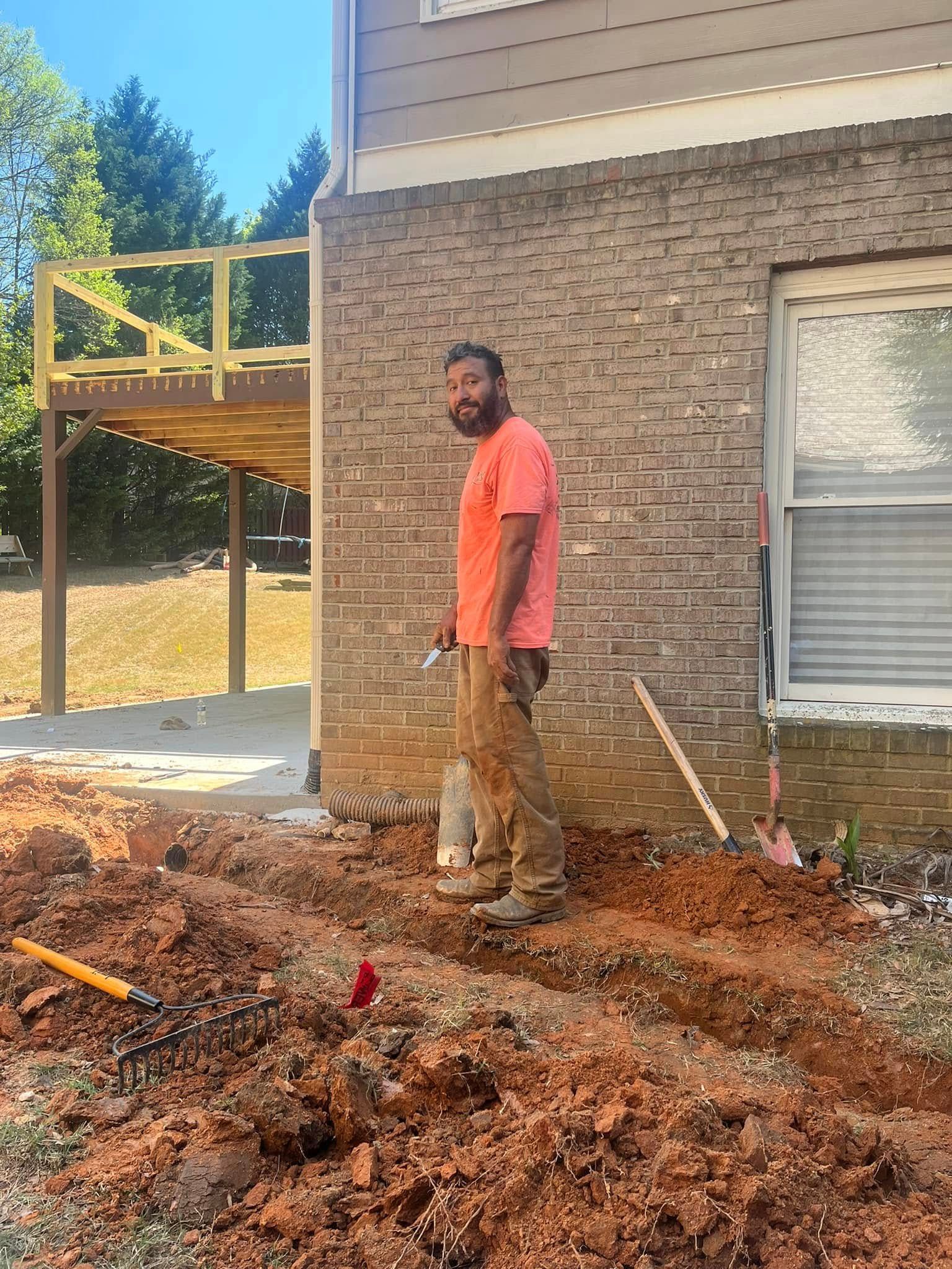Man standing by a brick house, digging in the dirt. A deck is visible in the background.