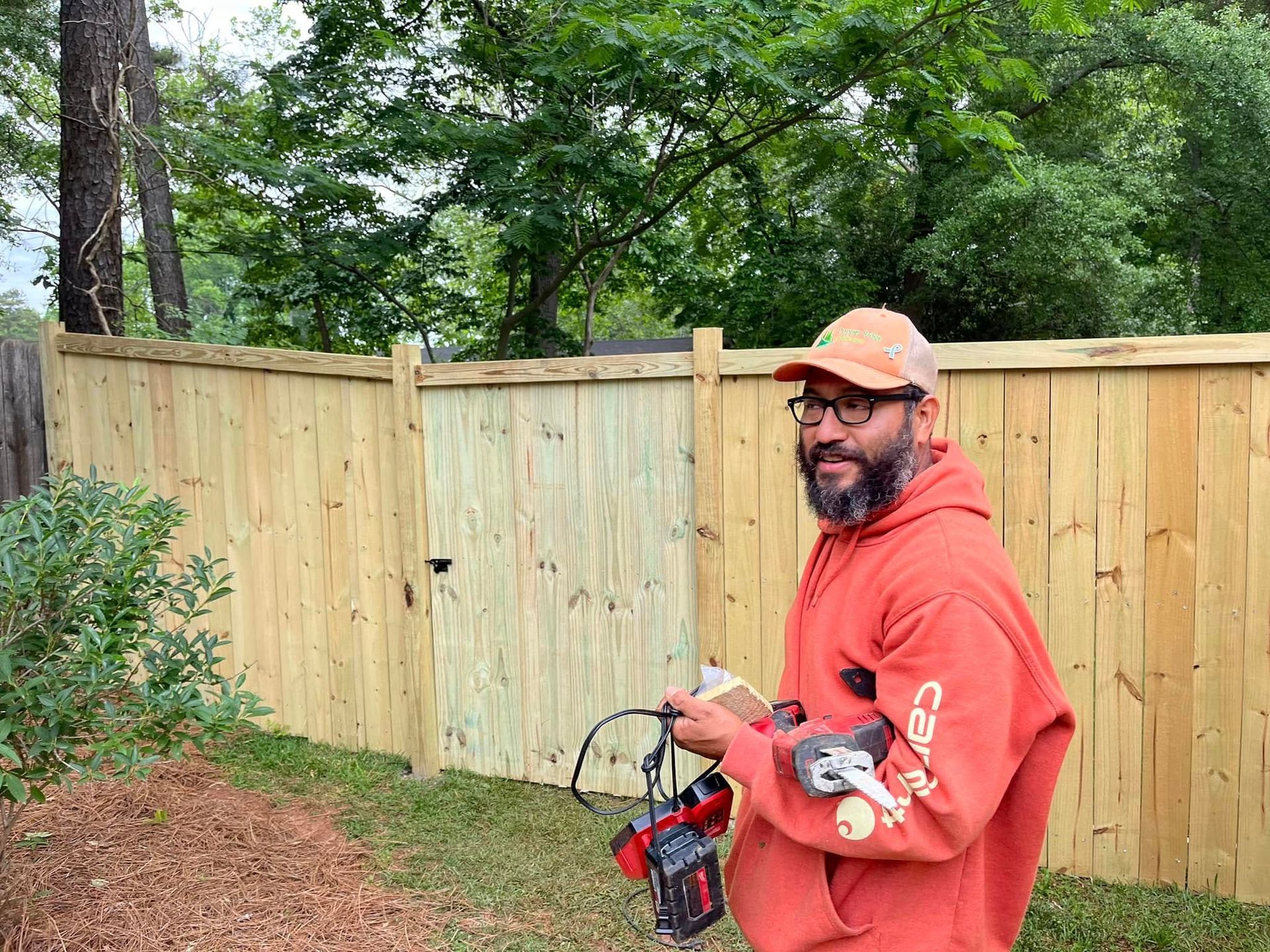 Man in orange hoodie stands in front of a new wooden fence, holding tools.