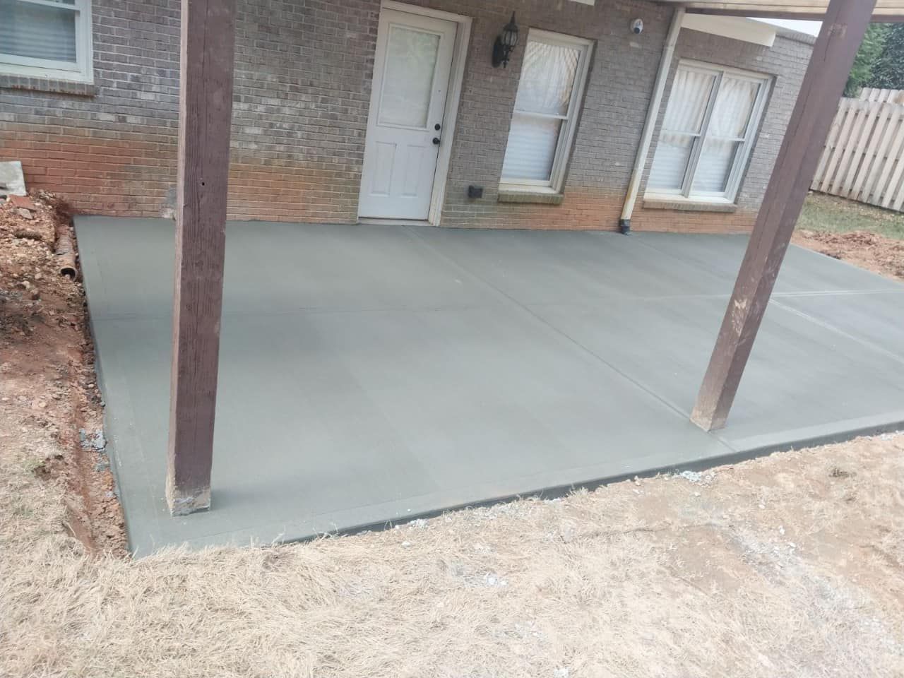 Newly poured concrete patio next to a brick house, under a wooden pergola.