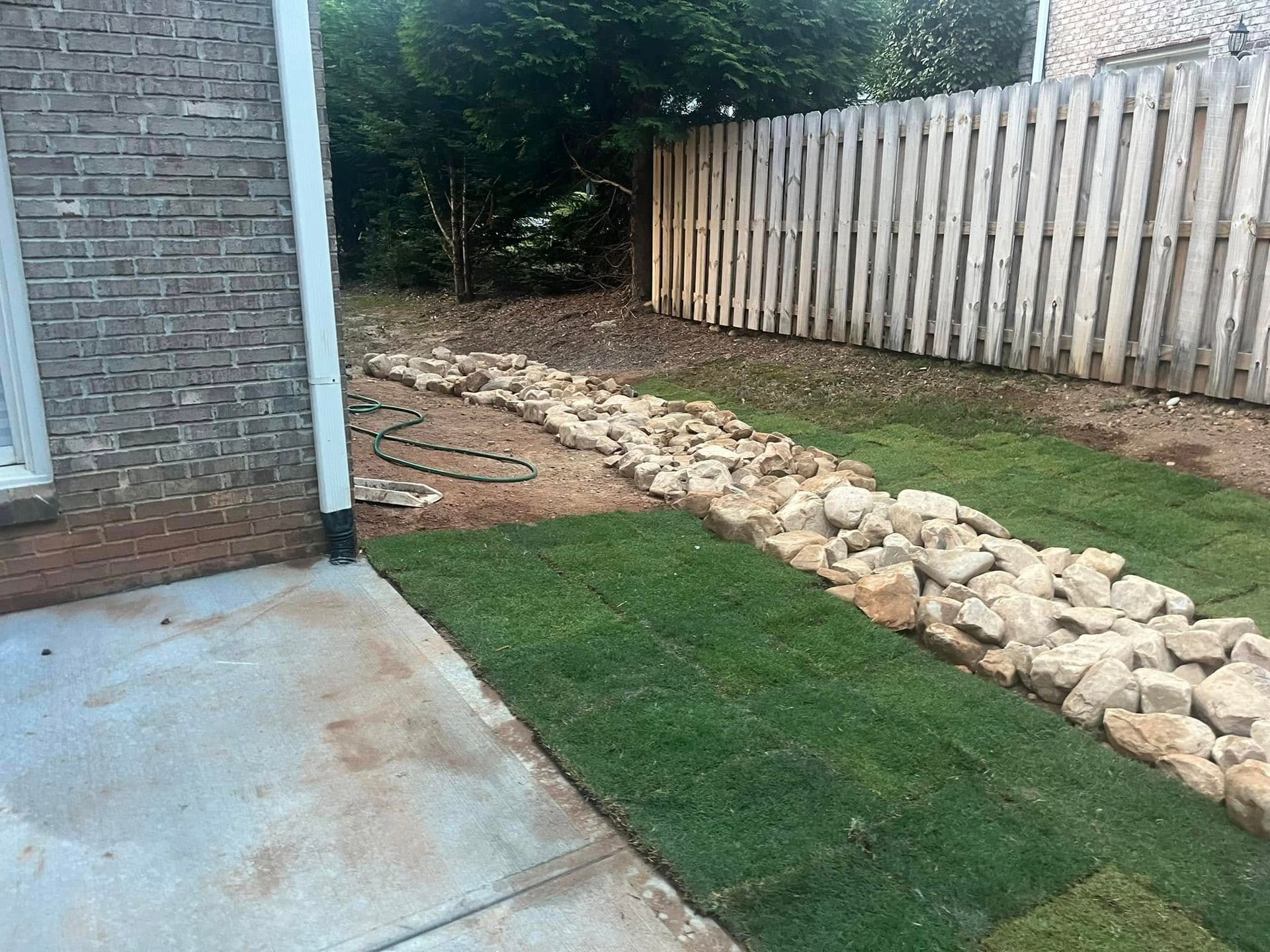 Newly laid sod alongside a rock-lined drainage channel next to a wooden fence and building.