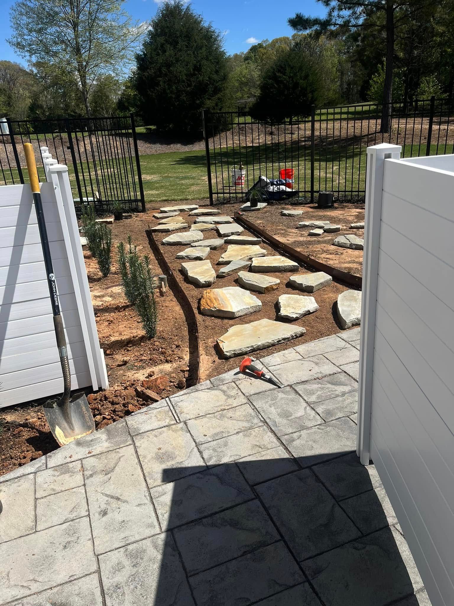 A stone pathway through a garden bed, between white fences, shovel, and plants.