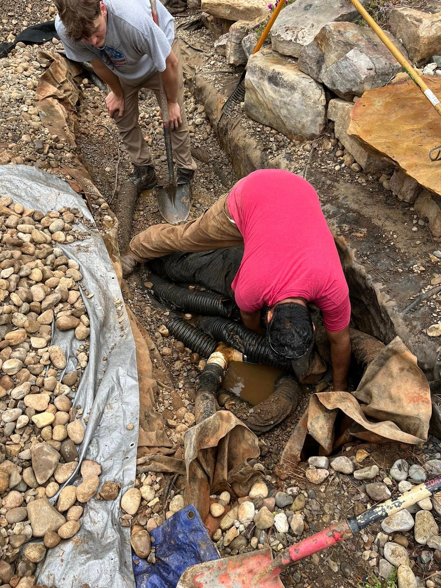 Two people work in a muddy trench. One in pink shirt leans down, the other standing uses a tool.