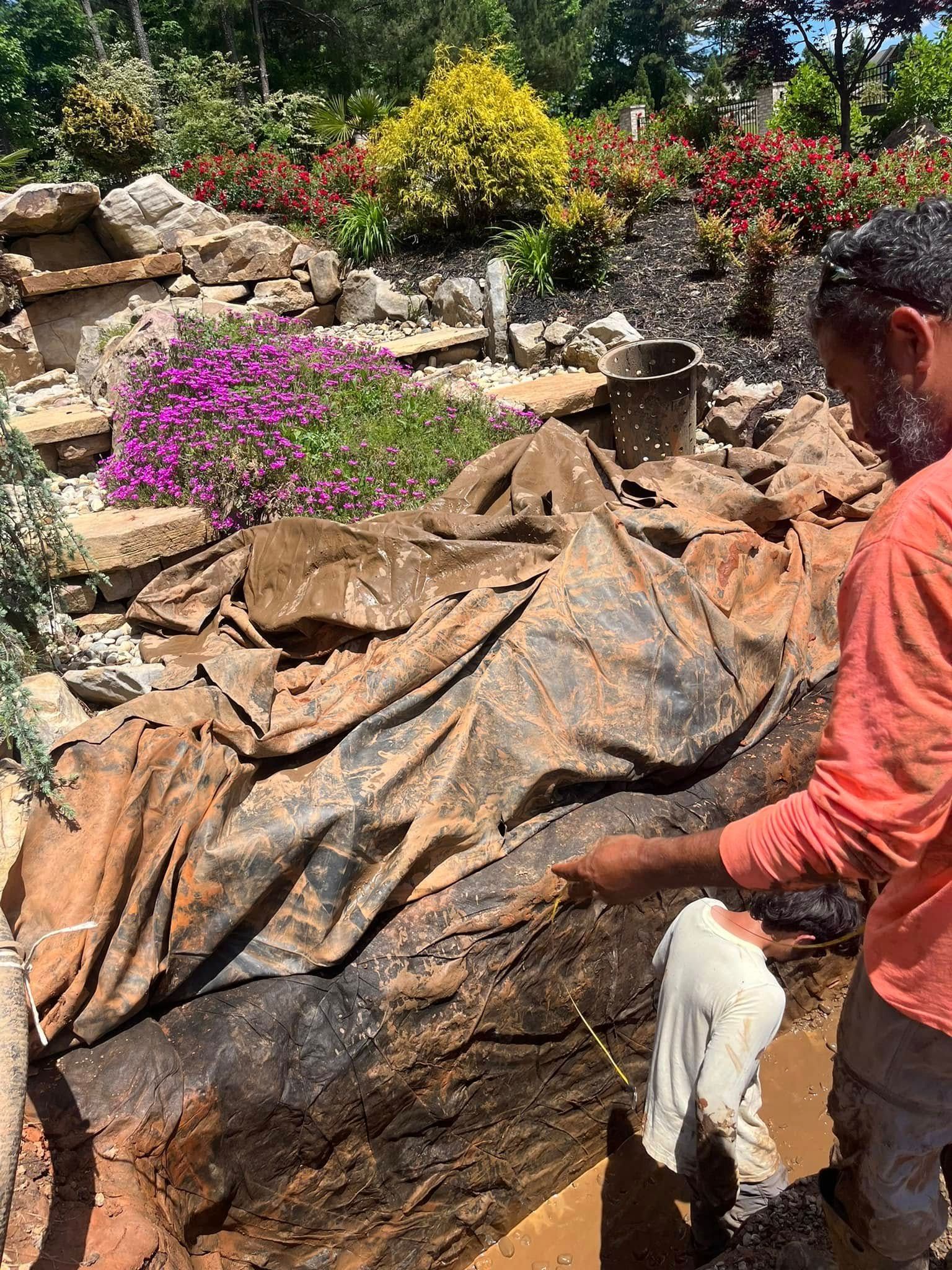 Man in orange shirt points at muddy ground covered with black tarp; another man is nearby.