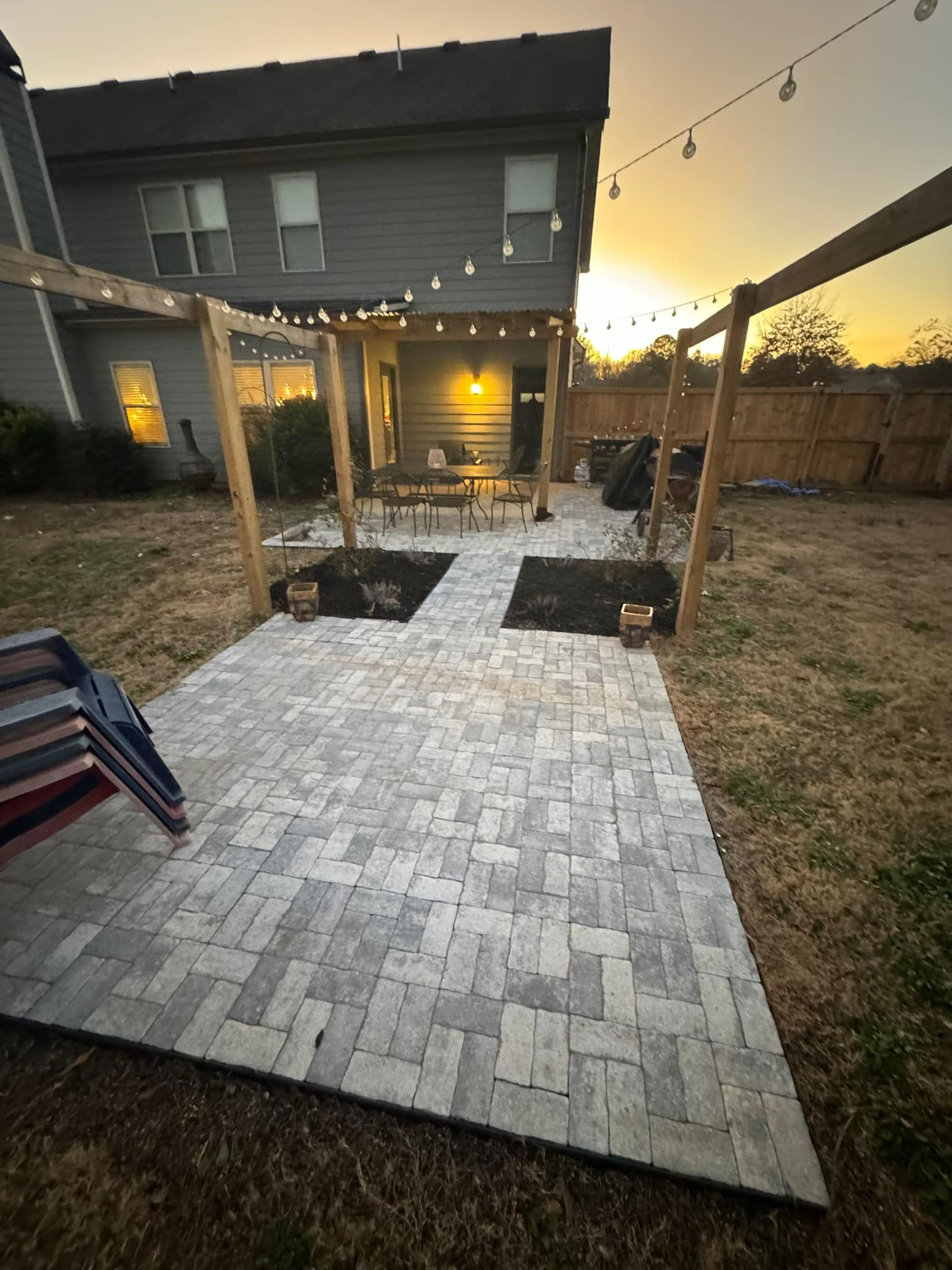 Backyard patio with brick pavers, string lights, pergola, and wooden fence.