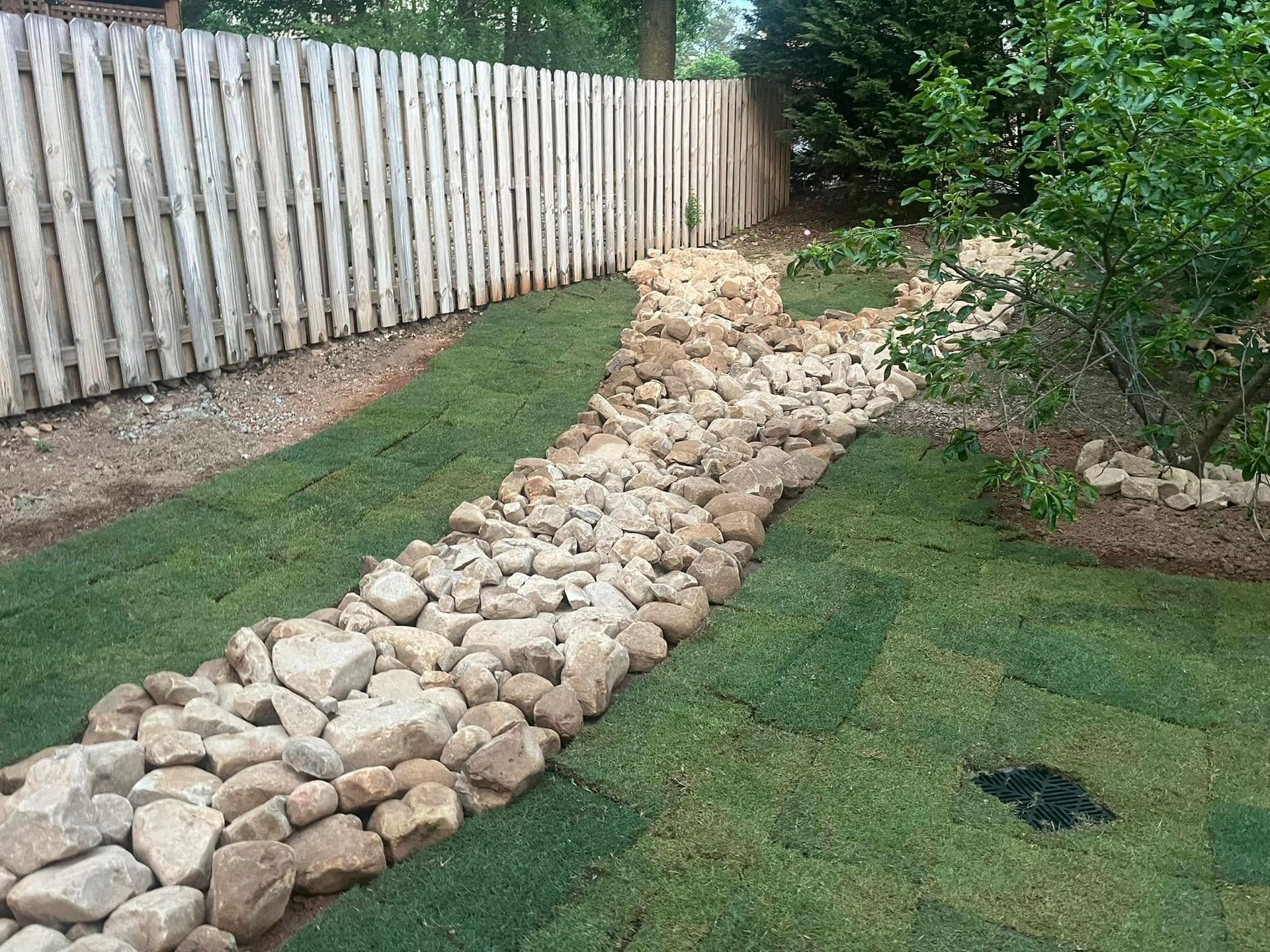 Rock-lined drainage channel in yard with new sod, beside a wooden fence and tree.
