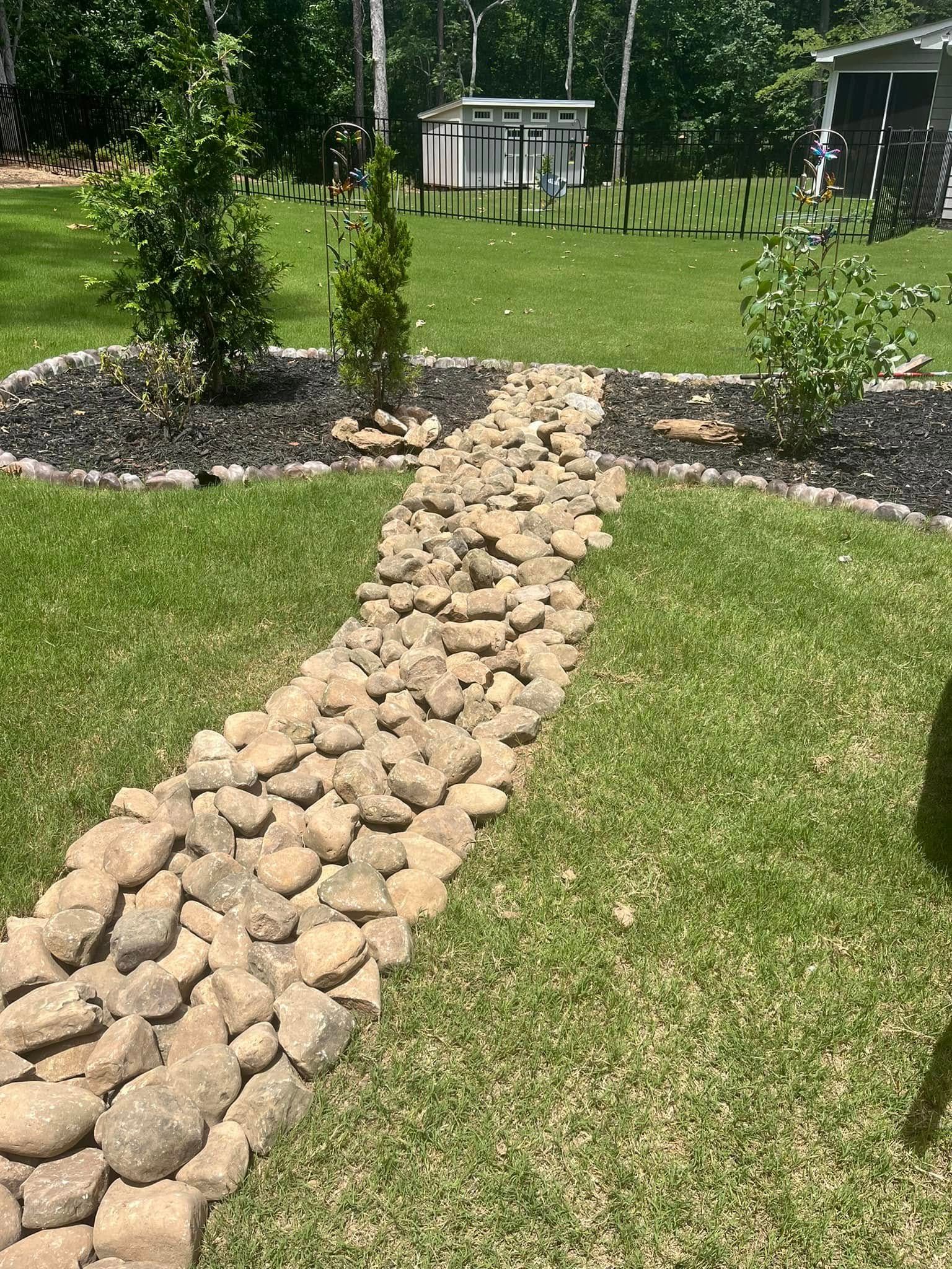 A rock-lined pathway through a grassy yard, with small trees and a shed in the background.