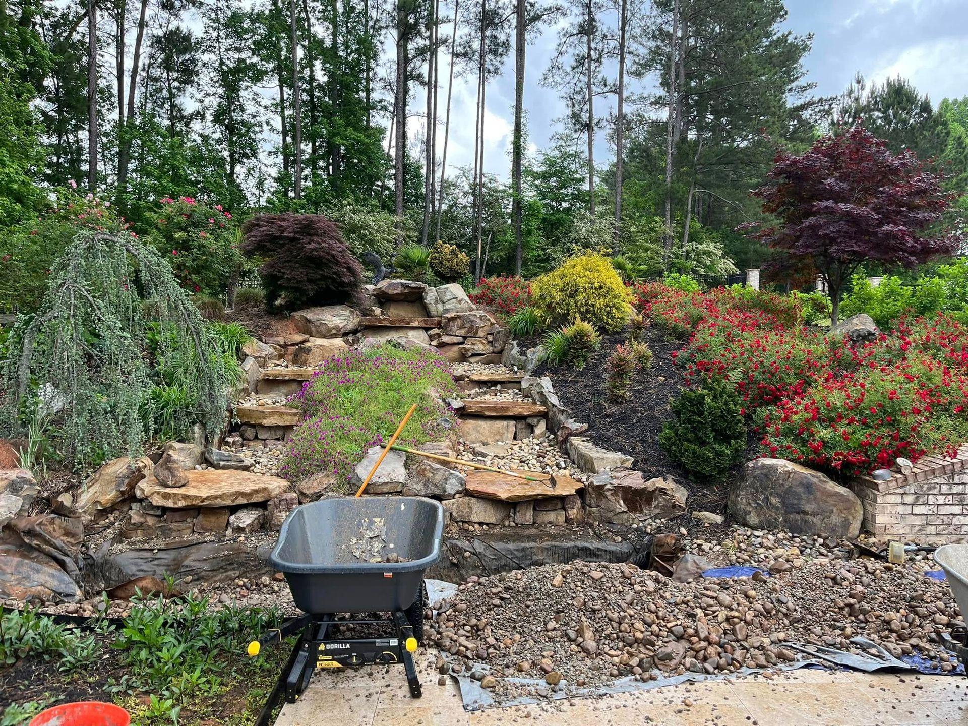 A stone tiered garden with a wheelbarrow filled with gravel, surrounded by colorful bushes and trees.