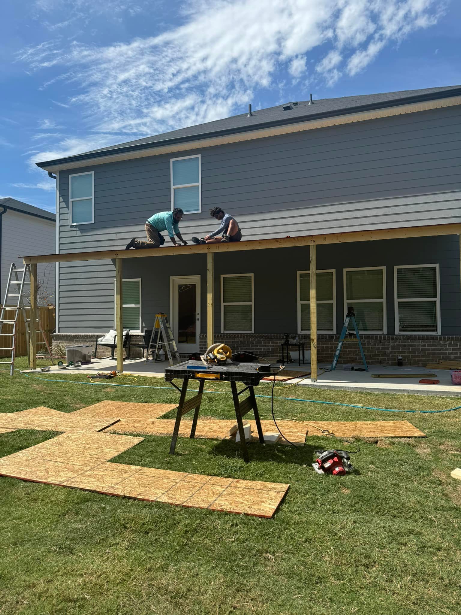 Two people building a deck attached to a two-story gray house. Saw table and wood scraps on grass in front.