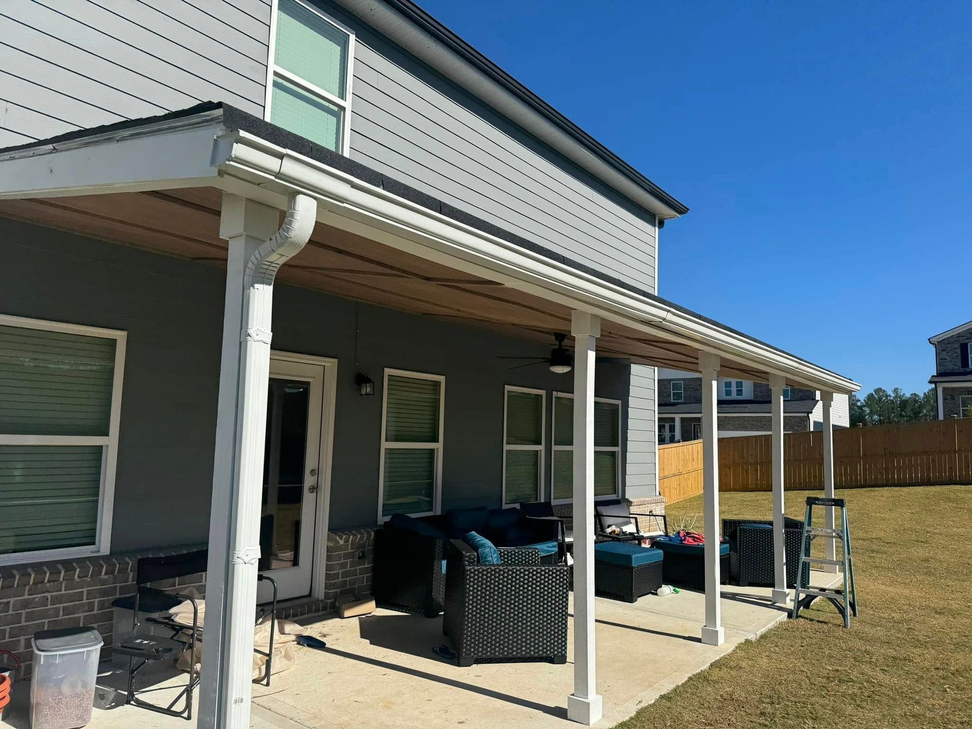 Backyard patio with white columns, gray siding, and a tan roof. Furnishings and lawn visible.