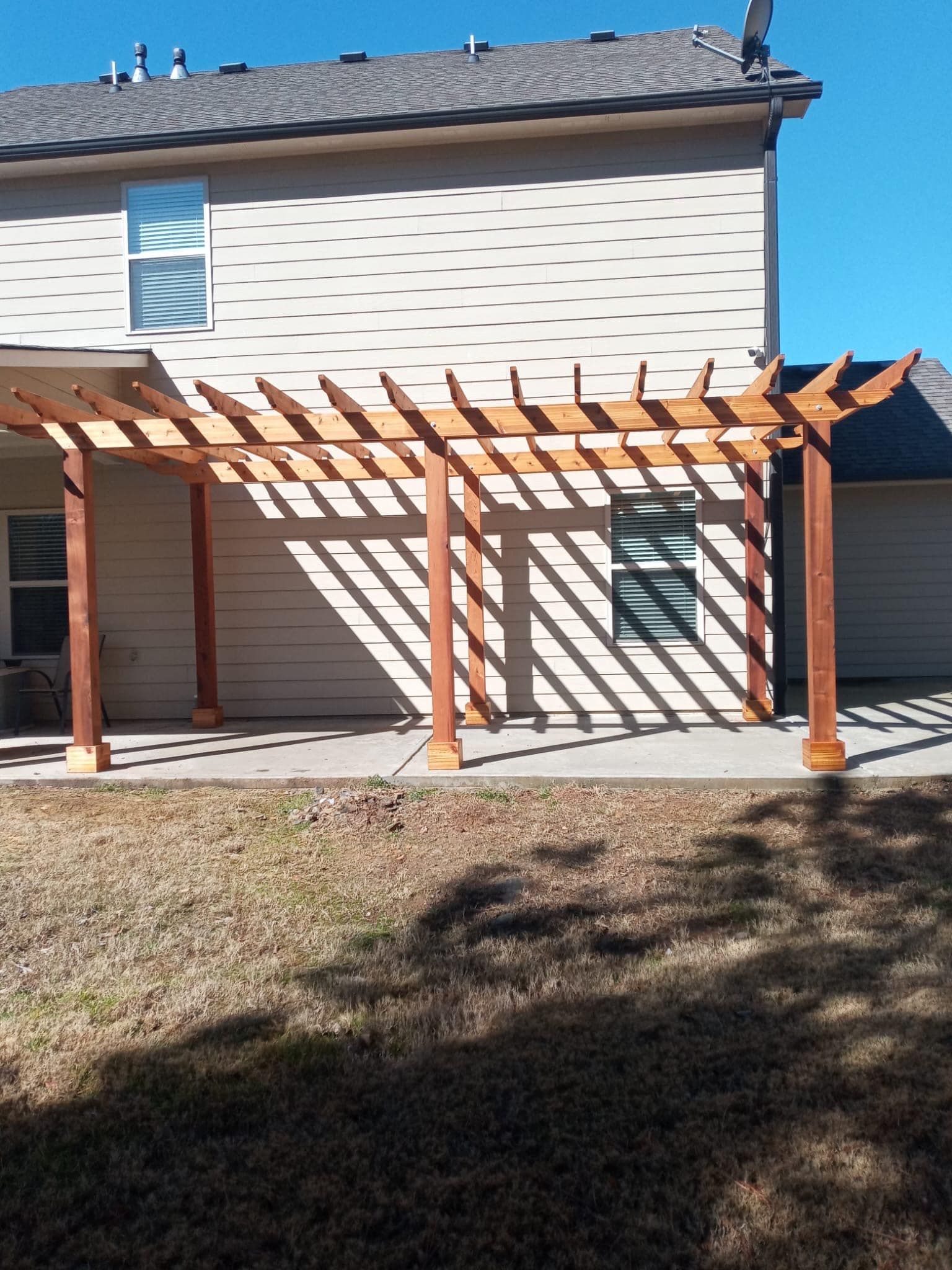 Wooden pergola attached to the back of a two-story beige house; shadows cast on the concrete patio.