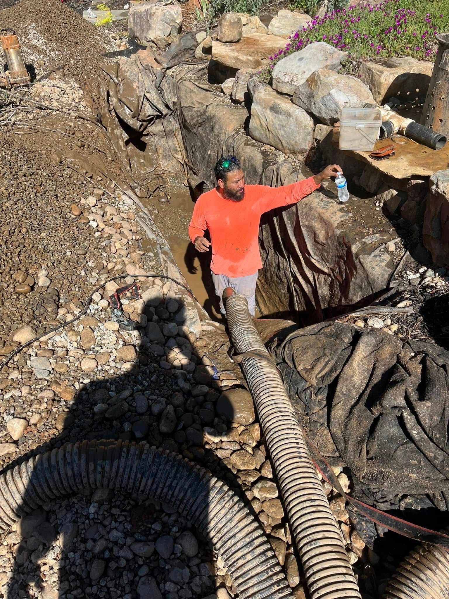 Man in orange shirt standing in a muddy ditch, holding a bottle. Surrounded by rocks, hose, and dark material.
