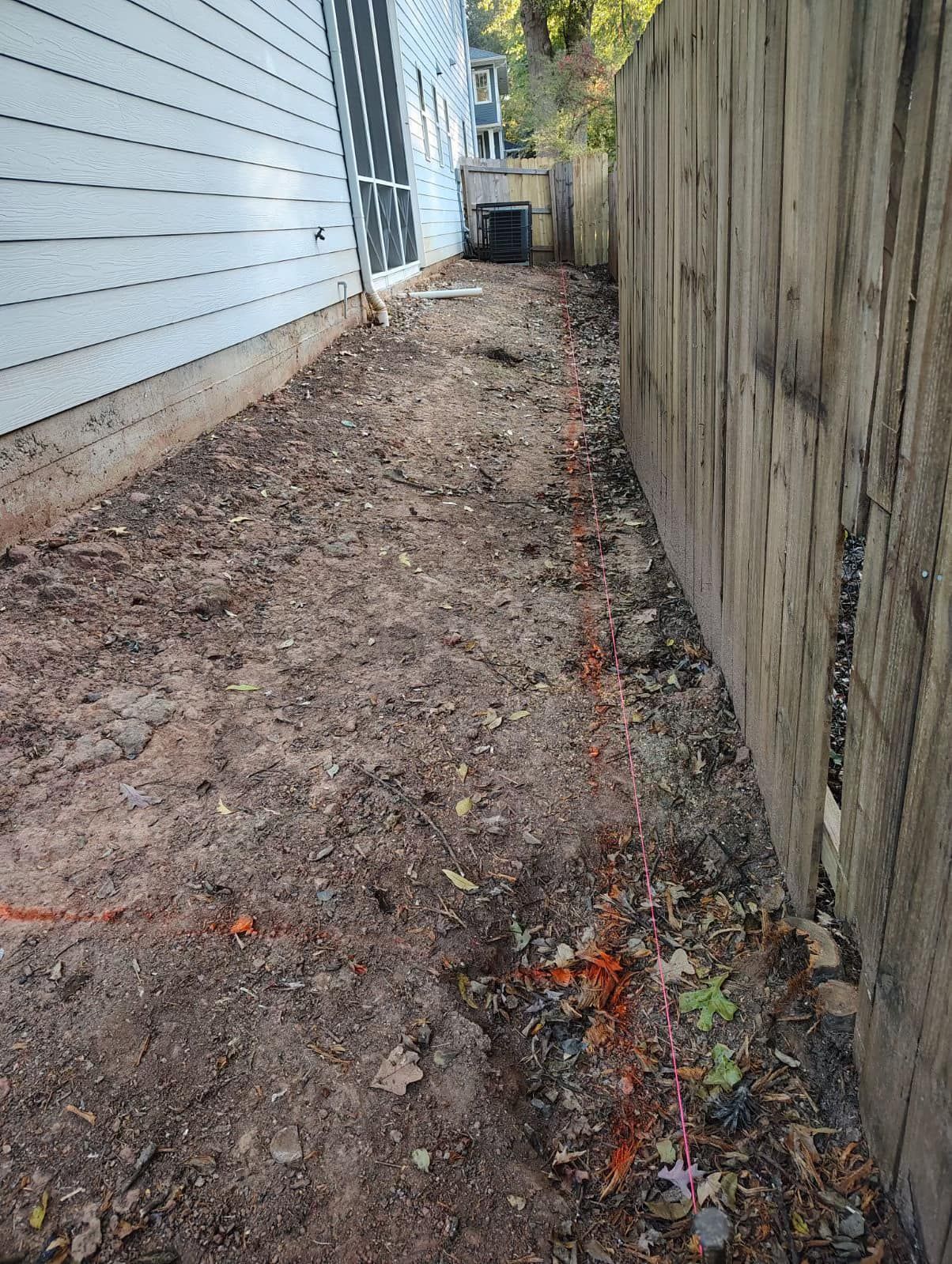 Narrow dirt path between a white-sided building and wooden fence, marked with orange paint.