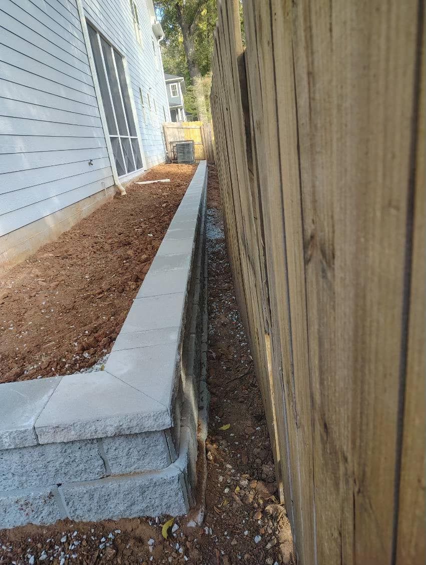 Retaining wall alongside a building and wooden fence. Brown dirt and gravel visible.