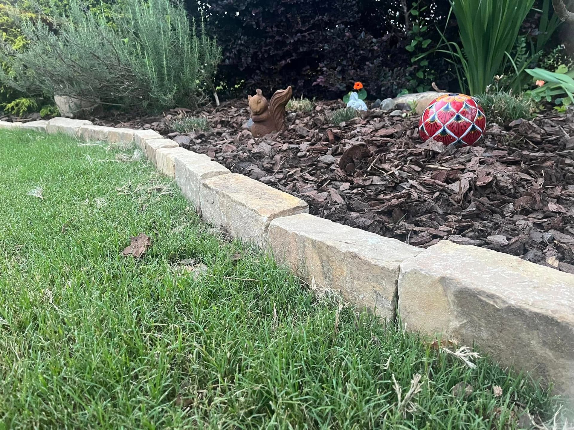 Stone border separating green grass from a garden bed with mulch and plants.