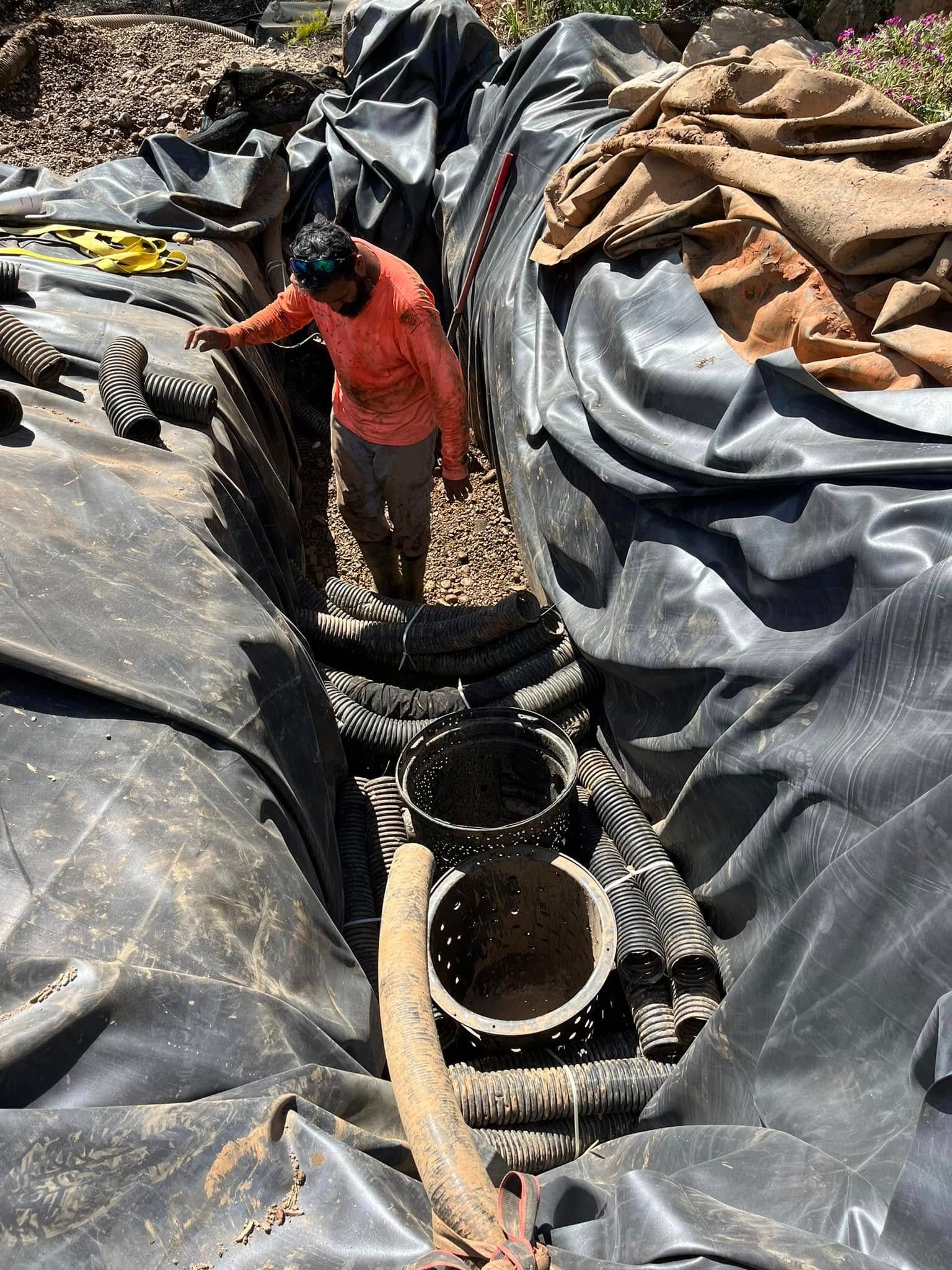 Person standing in lined trench with pipes; black liner, brown earth, and burlap visible.