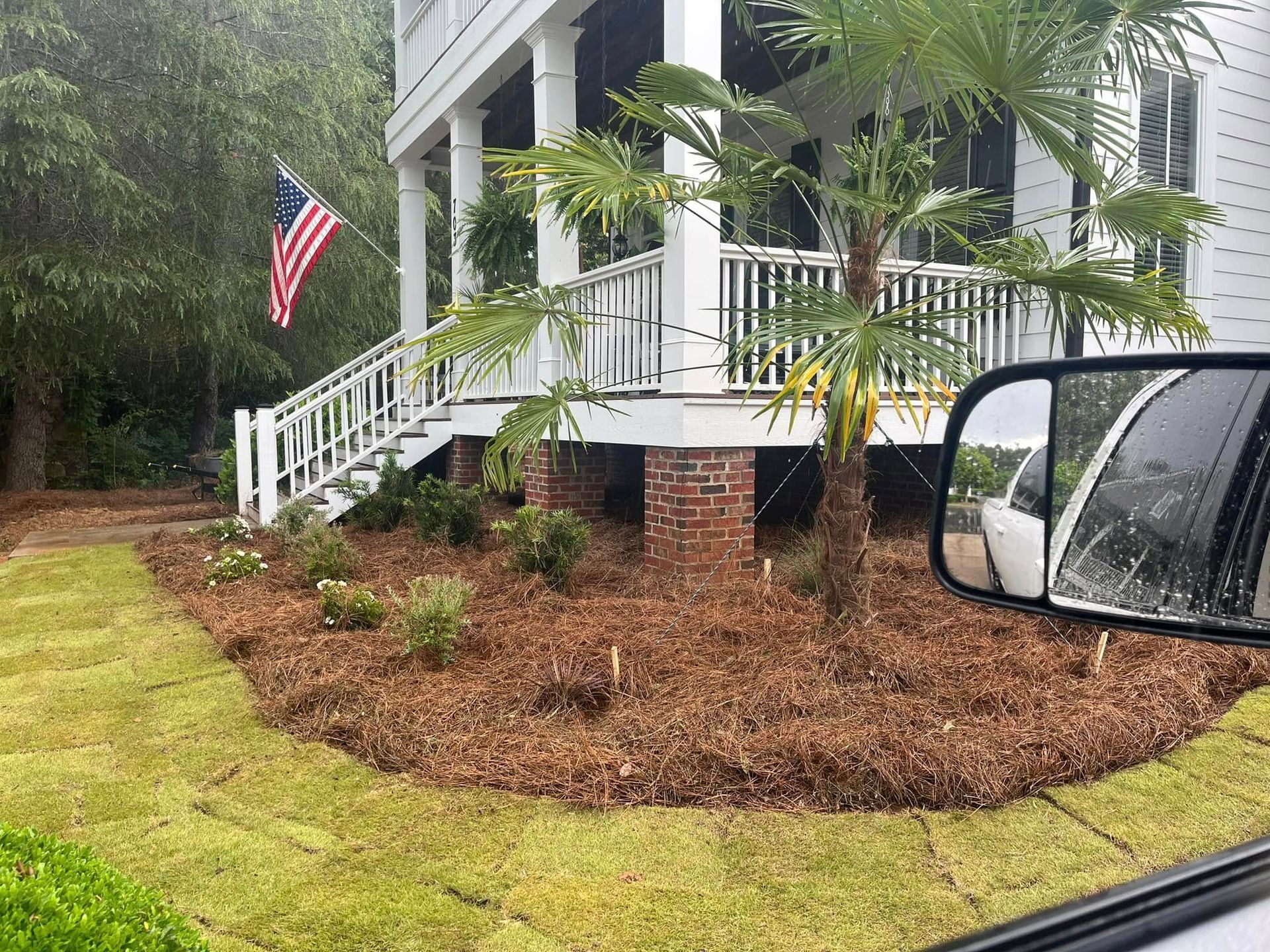 A house with a porch and American flag, with landscaping and a car side mirror in the foreground.