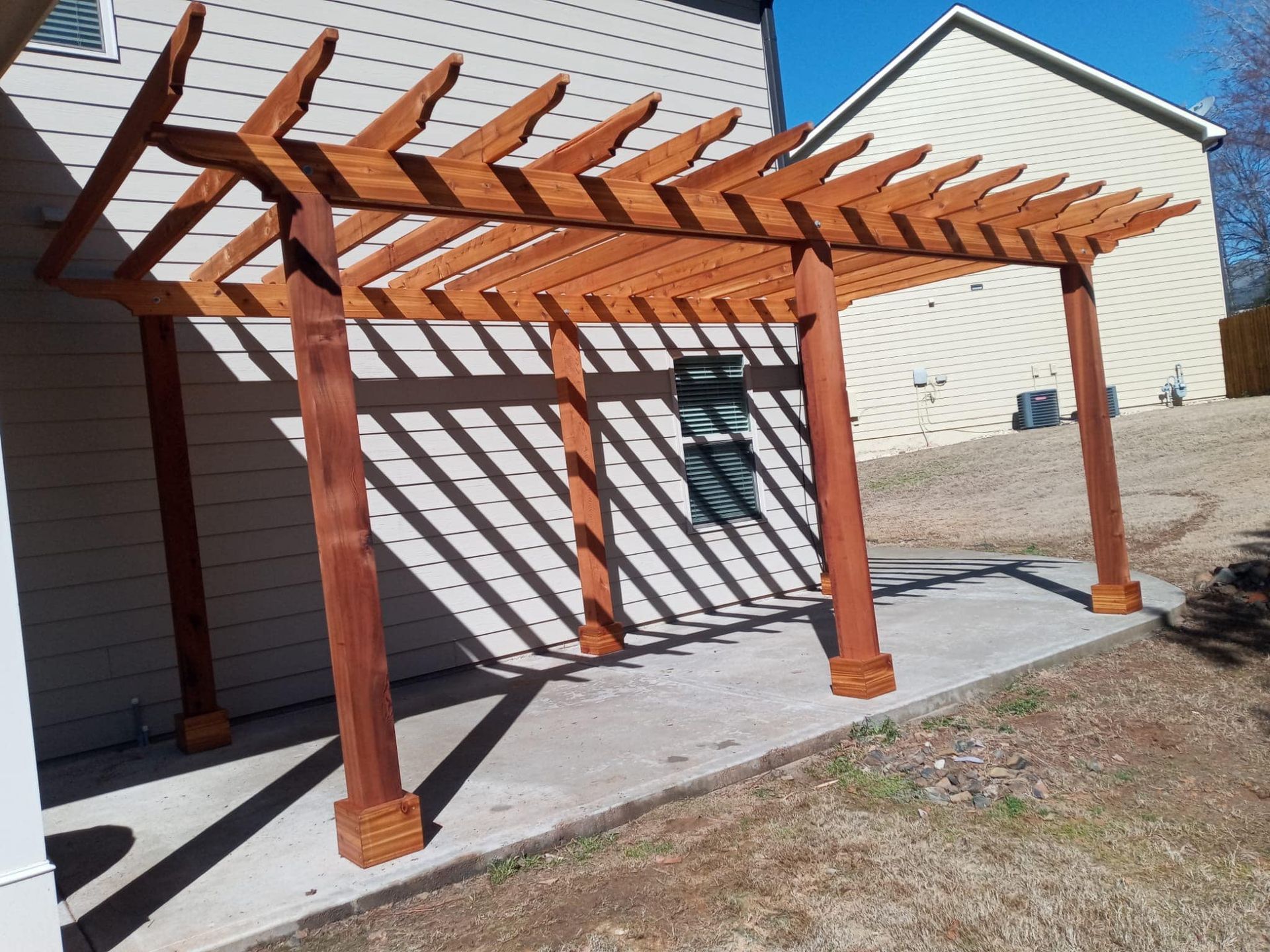 Wooden pergola attached to a light-colored house, casting shadows on a concrete patio in sunlight.