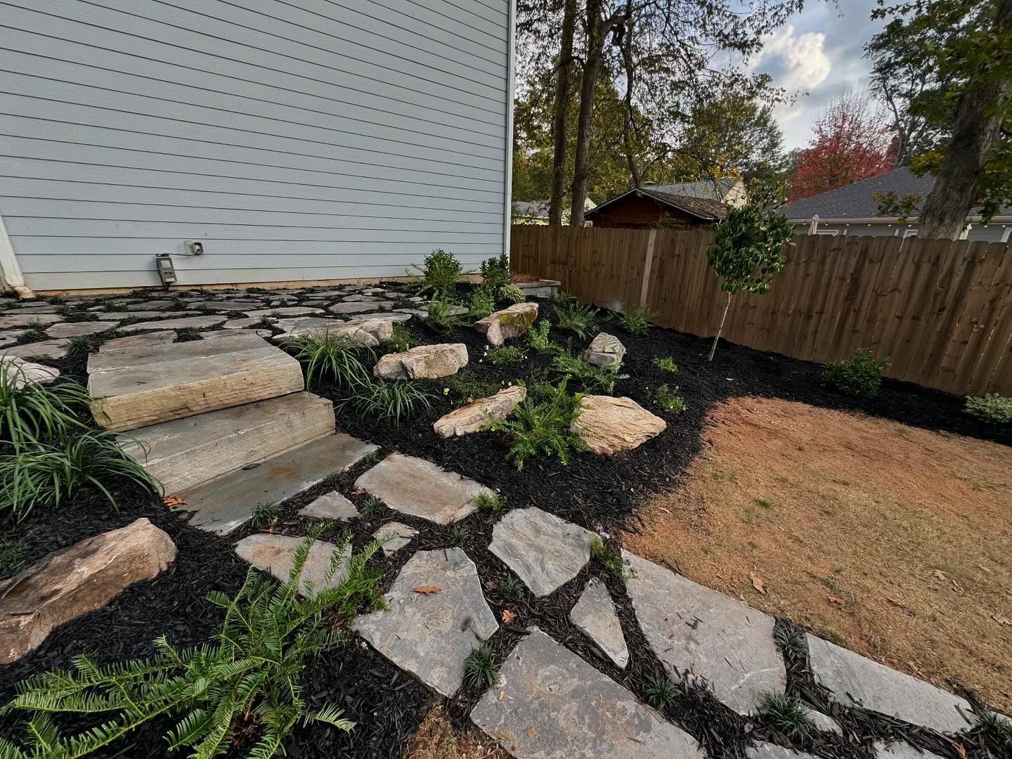 Stone pathway and steps in a yard with black mulch, plants, and a wooden fence.