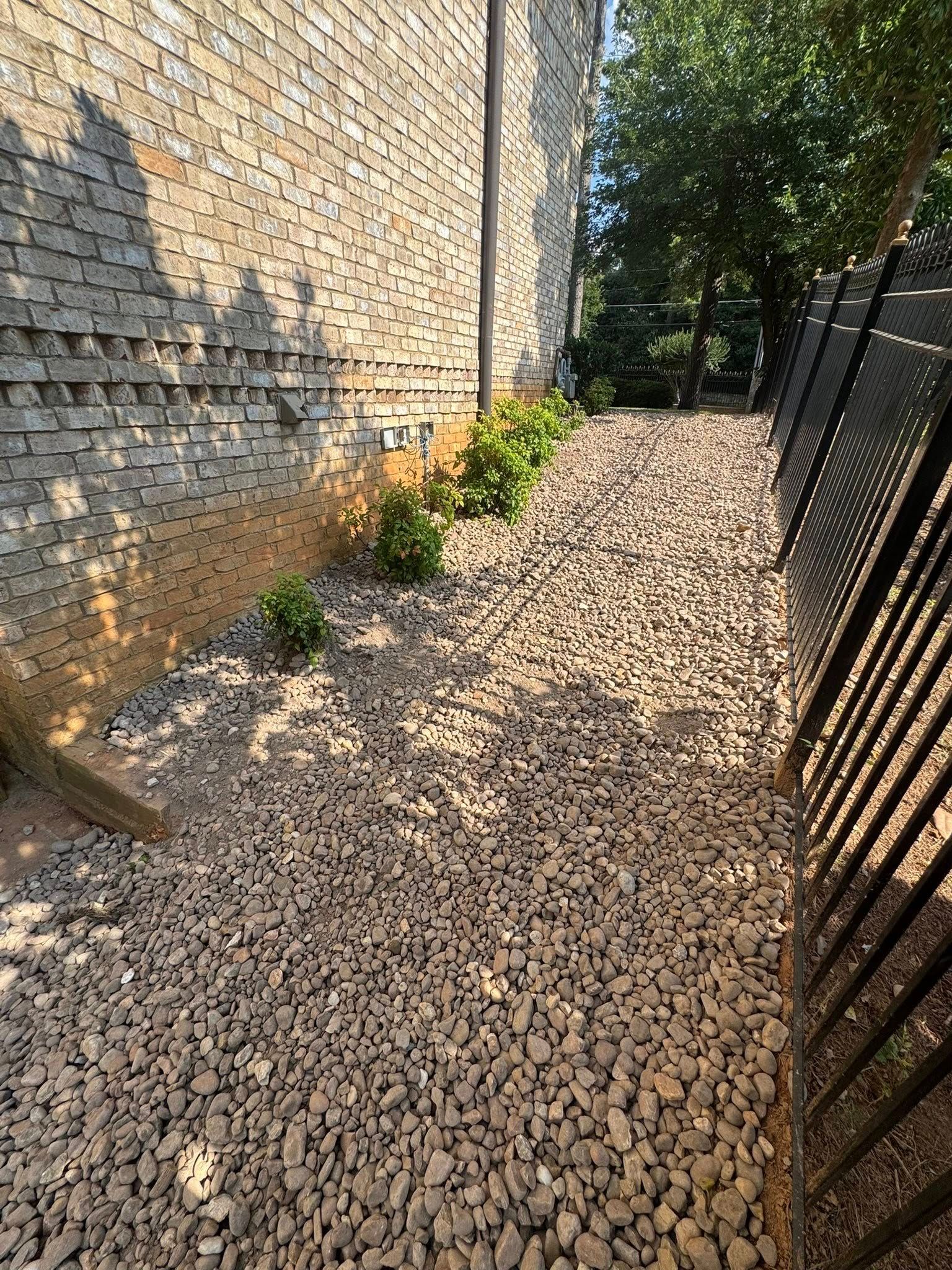 Gravel path beside a stone wall and black metal fence. Some small plants are growing along the wall.