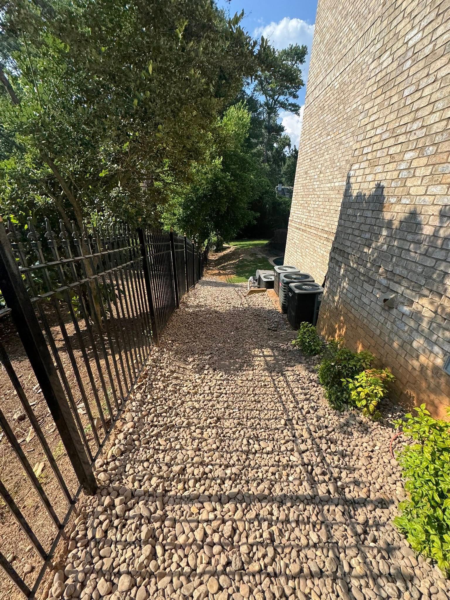 Gravel pathway beside a brick wall and black fence, with landscaping and utility boxes.
