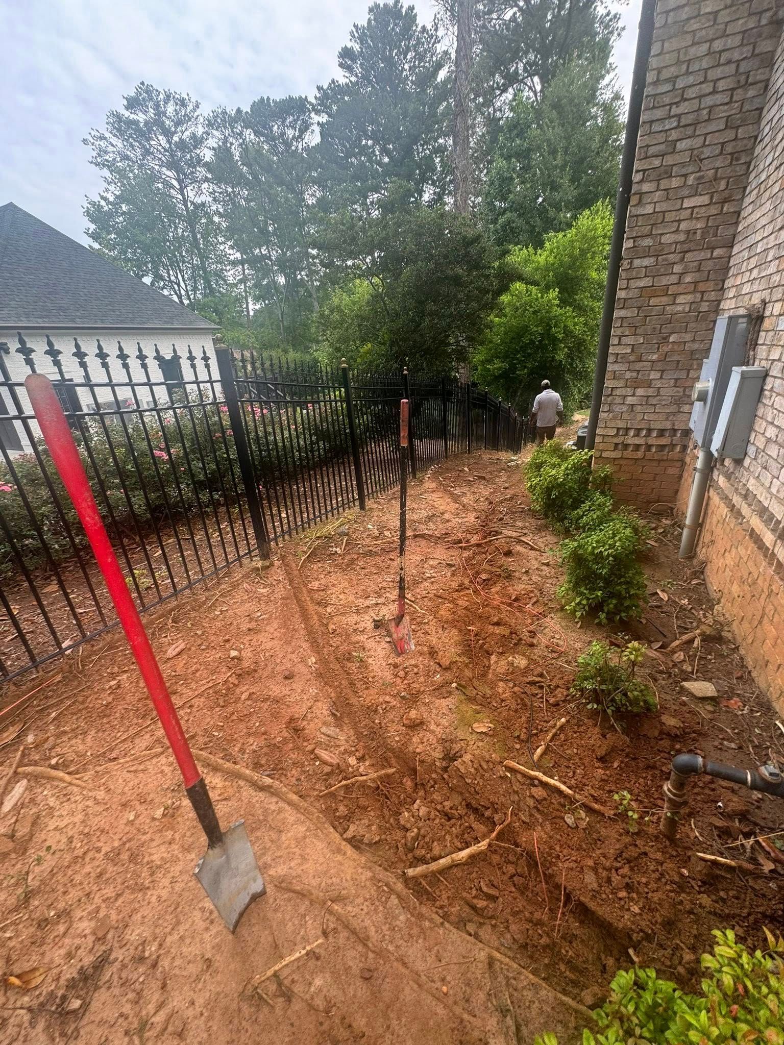 A shovel rests on the ground next to a trench. A black fence and brick building are visible in the background.