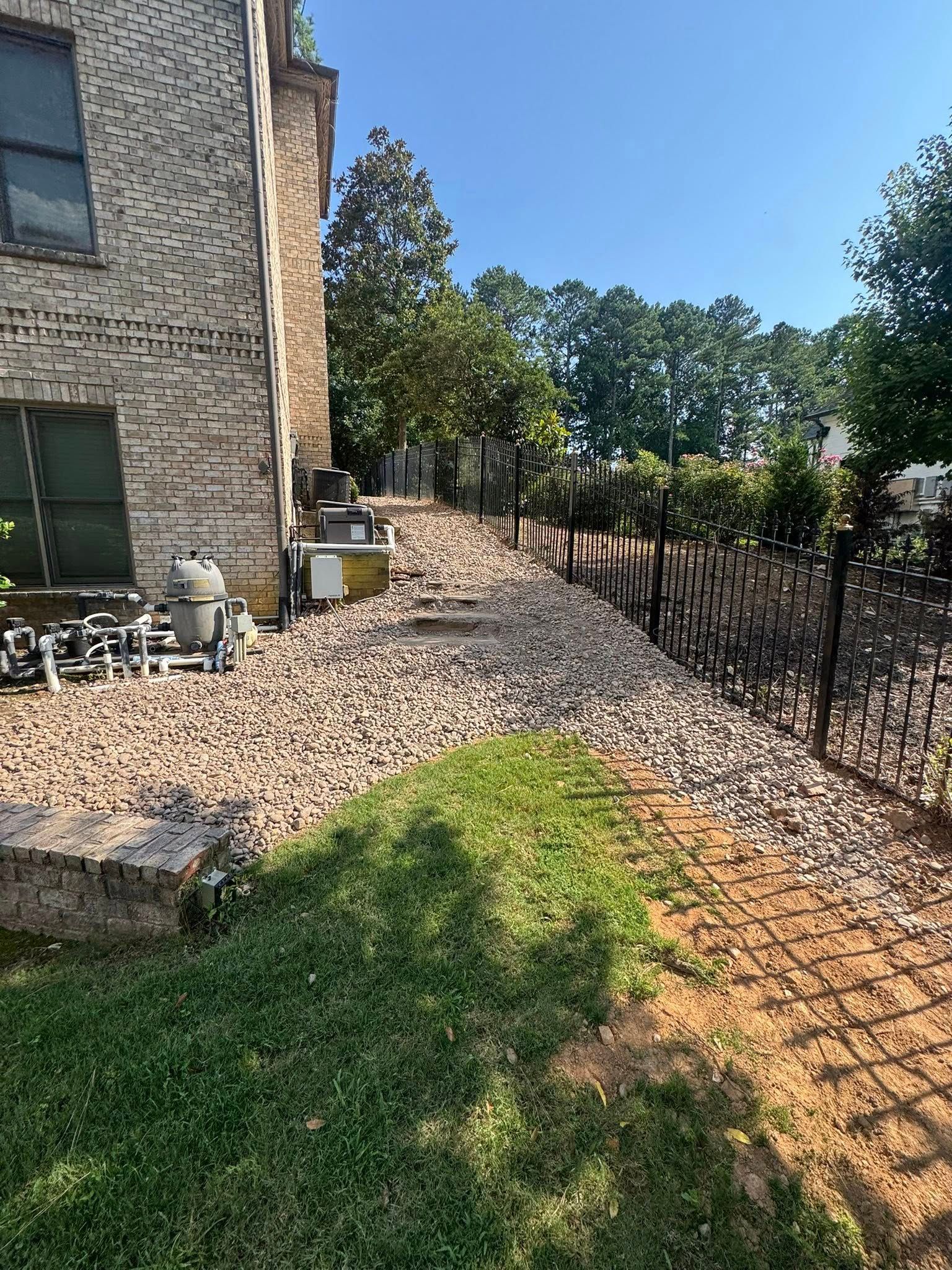 Gravel path along a brick building and decorative fence, with grass in the foreground and trees in the background under a blue sky.