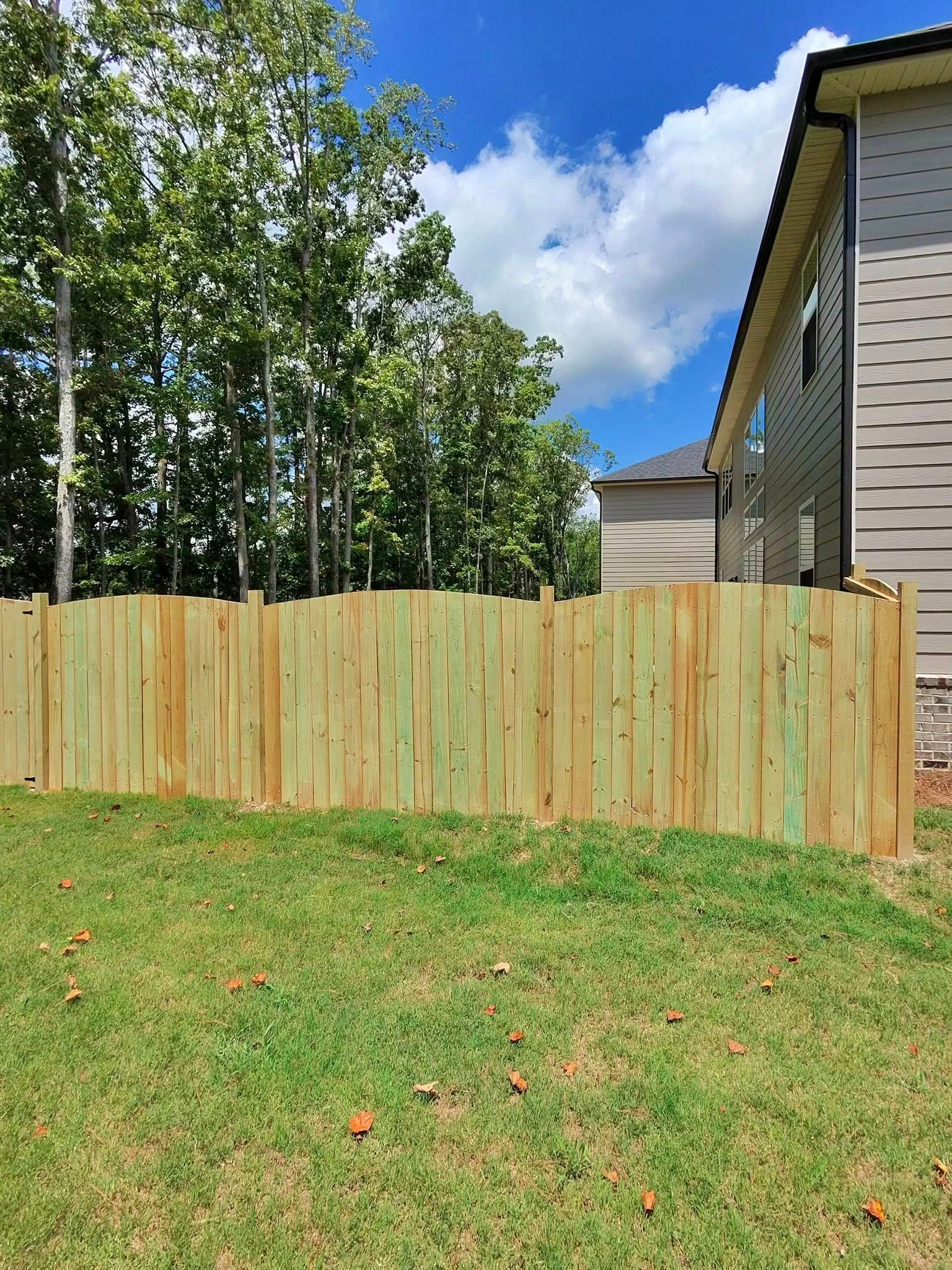 Wooden fence in a backyard with green grass, trees, and a house under a blue sky.