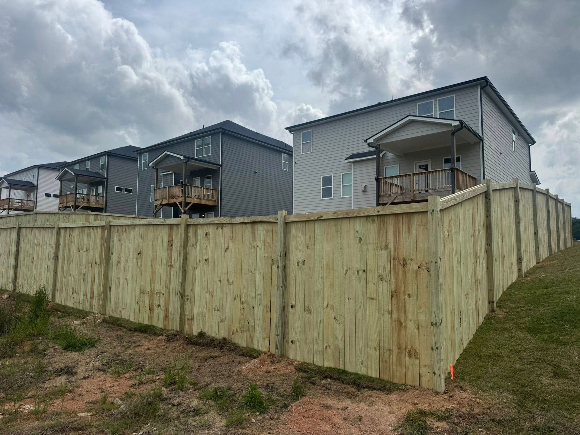 New wooden fence in front of several two-story gray houses with balconies under a cloudy sky.