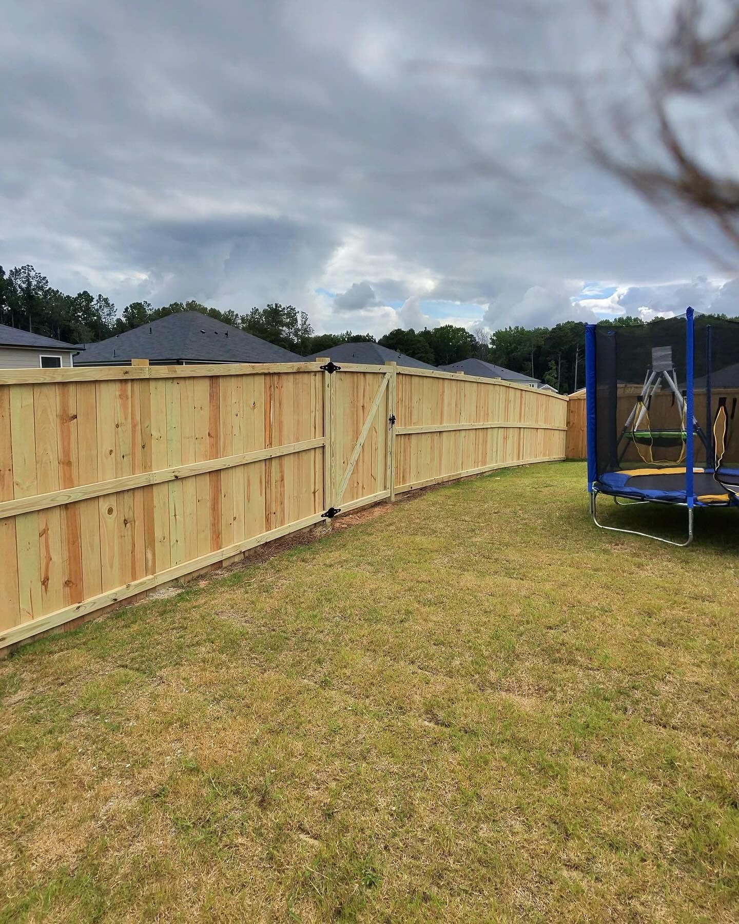 A wooden fence encloses a backyard with a trampoline under an overcast sky.