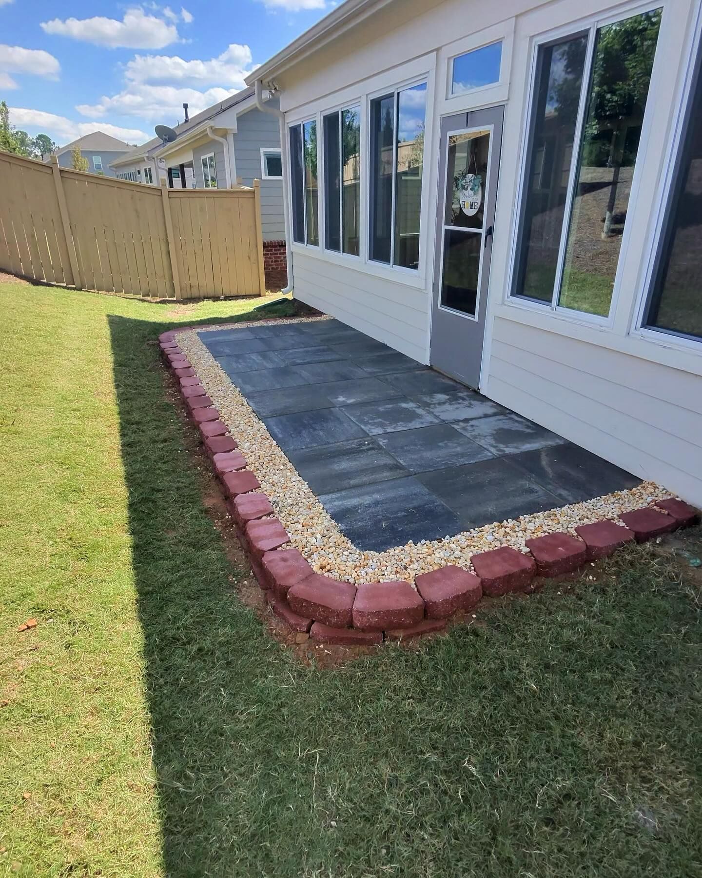Patio with gray stone tiles, red brick border, and gravel, next to a white house with windows and a door, on grass.