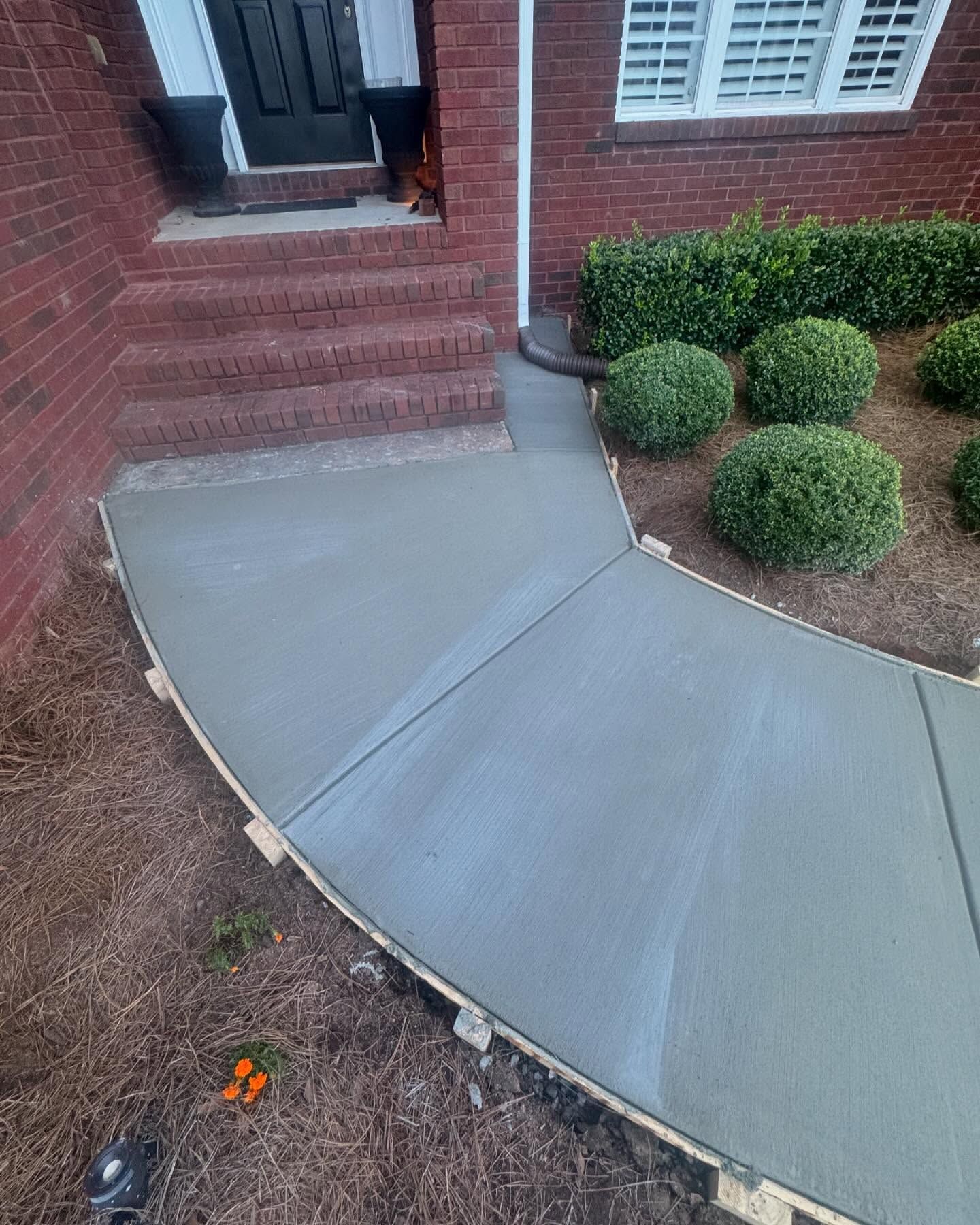 Newly poured concrete walkway curves toward a brick building, with stairs and bushes visible.