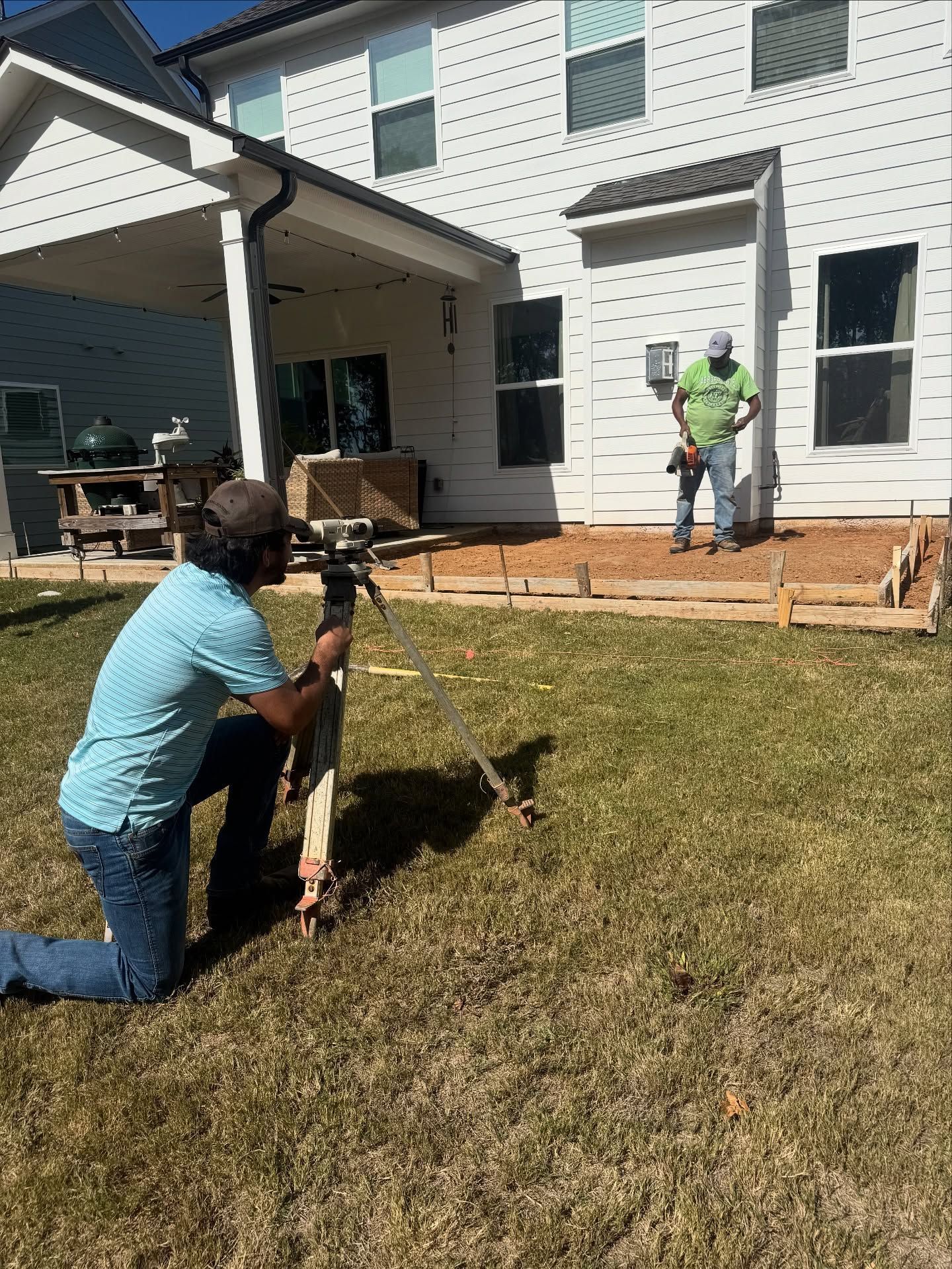Man kneels with tripod, leveling land for patio. Another man stands holding tools. Backyard.