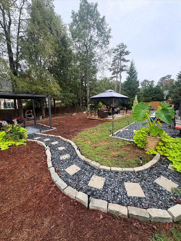 Backyard garden with stone path, mulch, and gazebo surrounded by trees.
