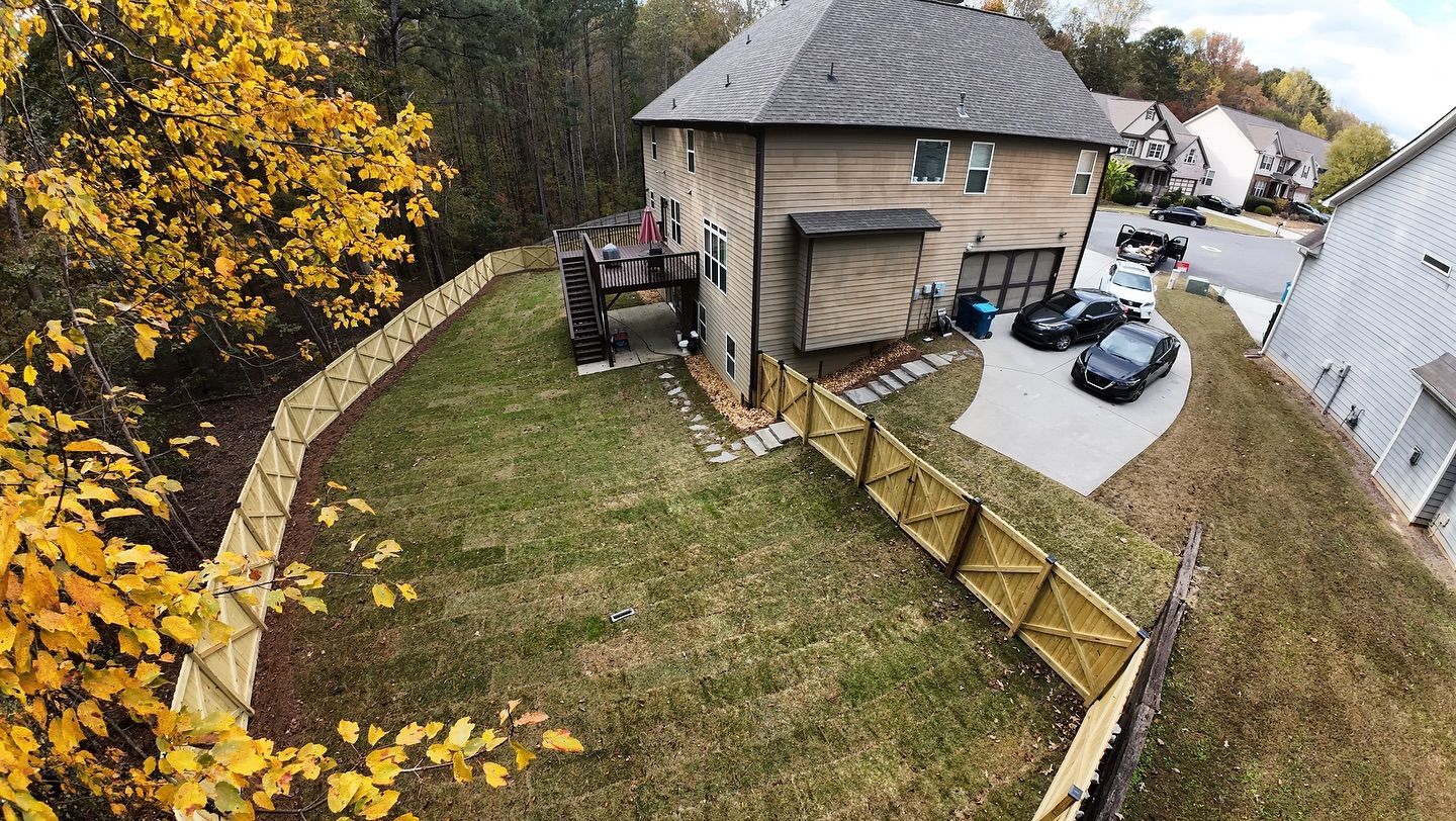 Aerial view of a two-story house with a wooden fence around the yard. Cars parked in driveway. Fall foliage.