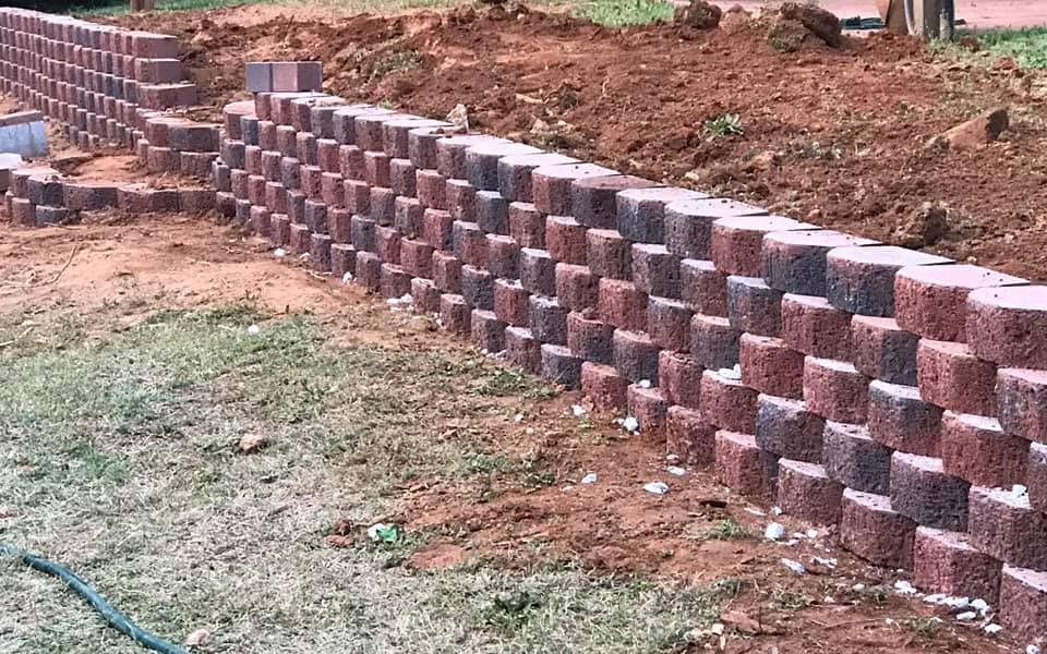 Red brick retaining wall being built on a grassy lawn with dirt and landscaping in the background.