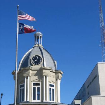 Courthouse with clock tower and flags of the United States and Texas against a blue sky.