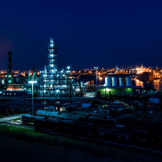 Night view of an industrial complex lit by numerous lights; train cars in the foreground, with dark blue sky.