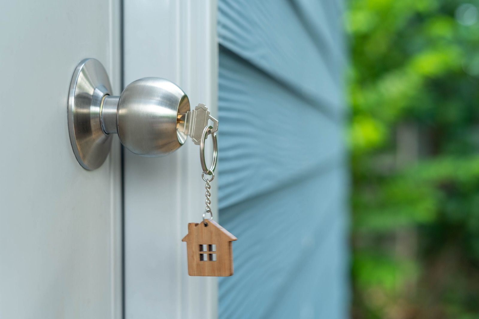 Key in door, house-shaped keychain hanging. Door is white, siding blue, with a blurred green background.