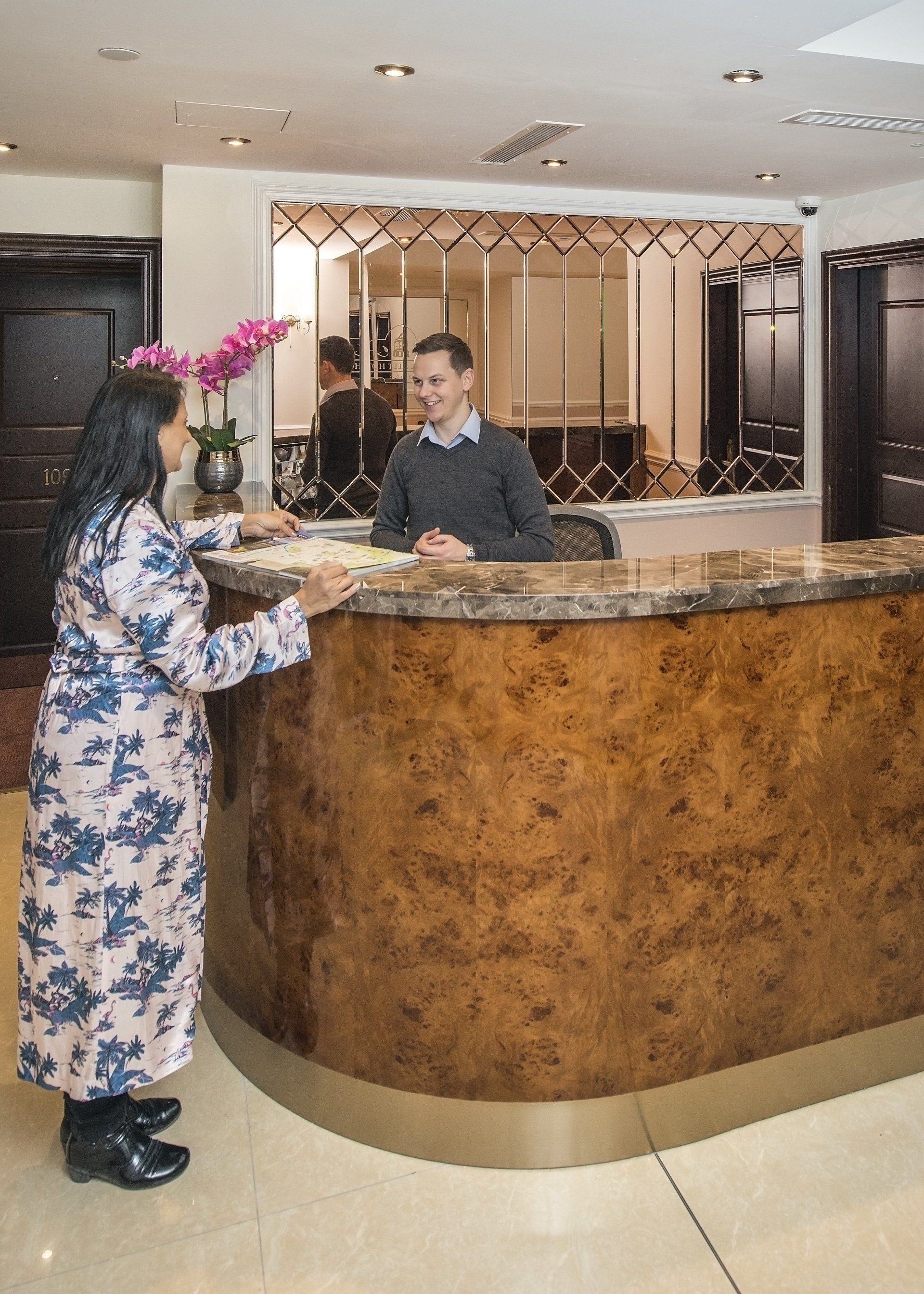 Woman at a hotel reception desk, talking to the clerk. Ornate wood desk and mirrored wall in the background.
