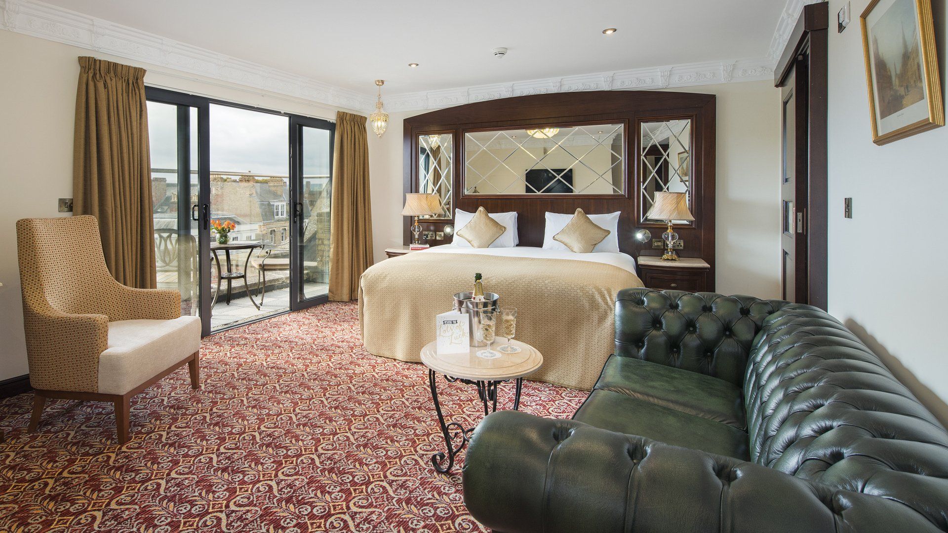 Hotel room with bed, balcony, sofa, and armchair; beige and green tones, ornate decor.