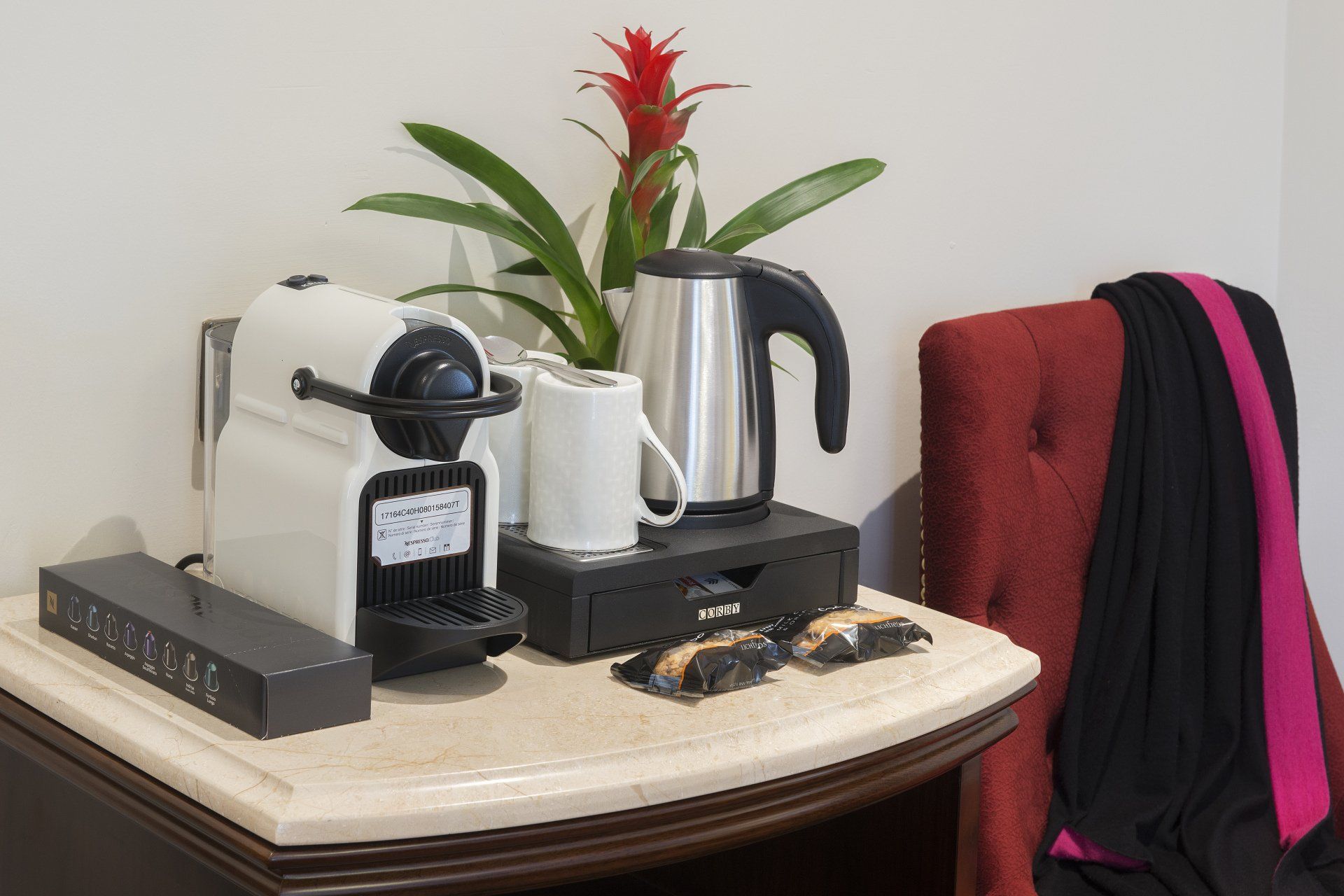 A coffee station on a table includes a coffee maker, electric kettle, mug, snacks, and a plant.