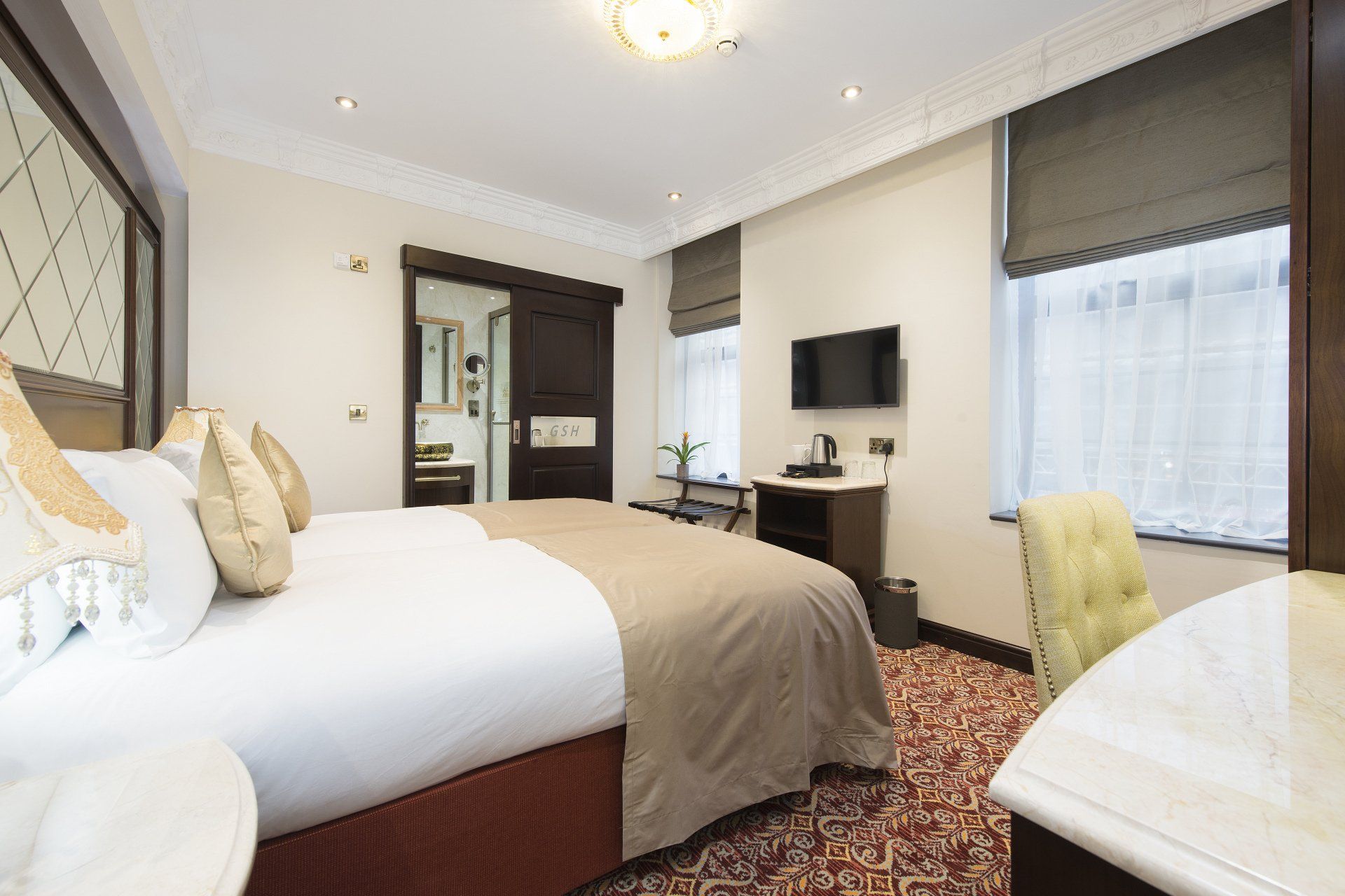 Hotel room: King-size bed with white bedding, TV on wall, dark wood door, window with shades, patterned carpet.