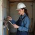 Woman in hard hat and overalls inspects plumbing, holding a tablet, indoors.