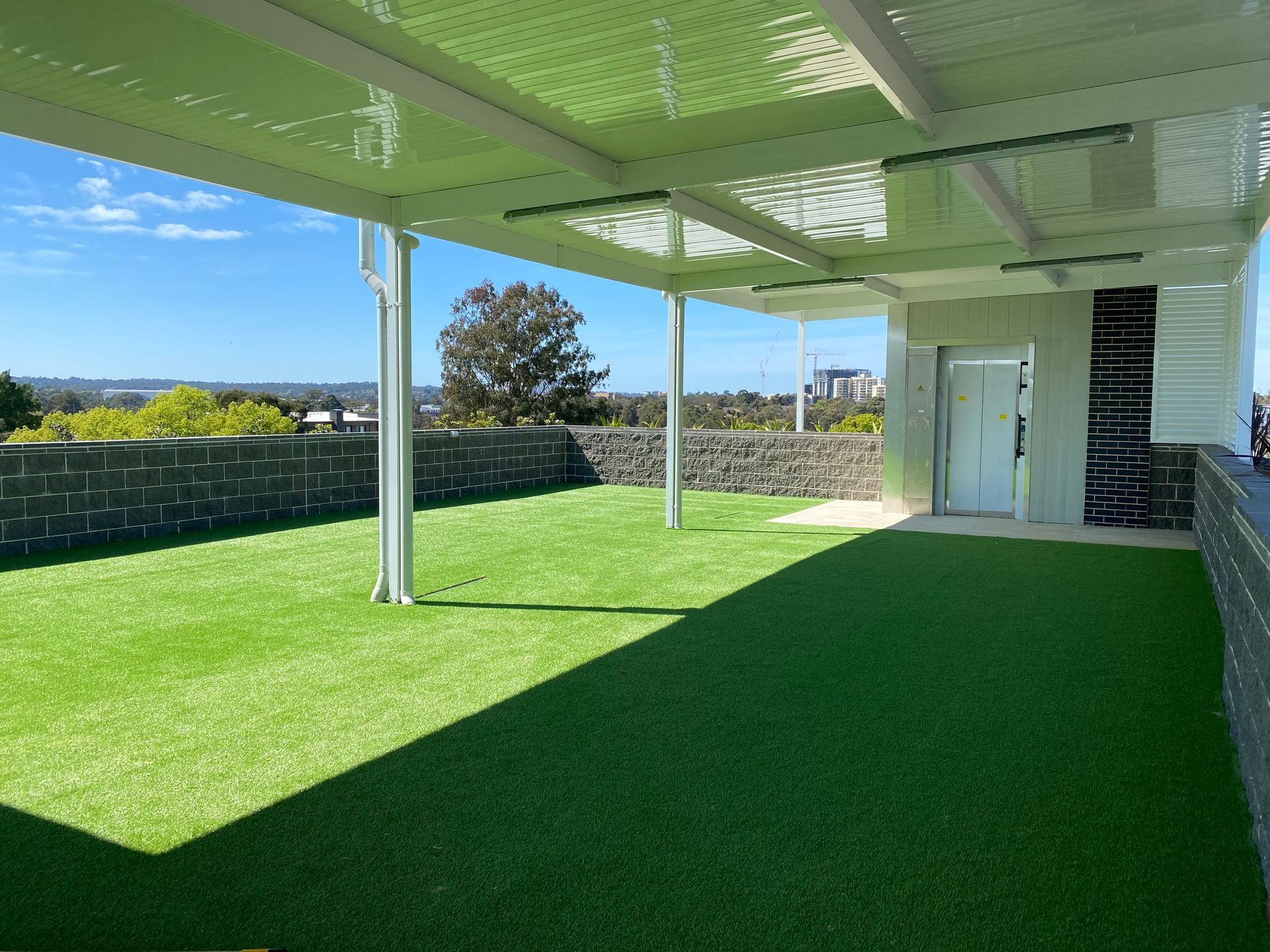Green artificial grass patio with a white covered roof. Blue sky in the background, a building to the right.