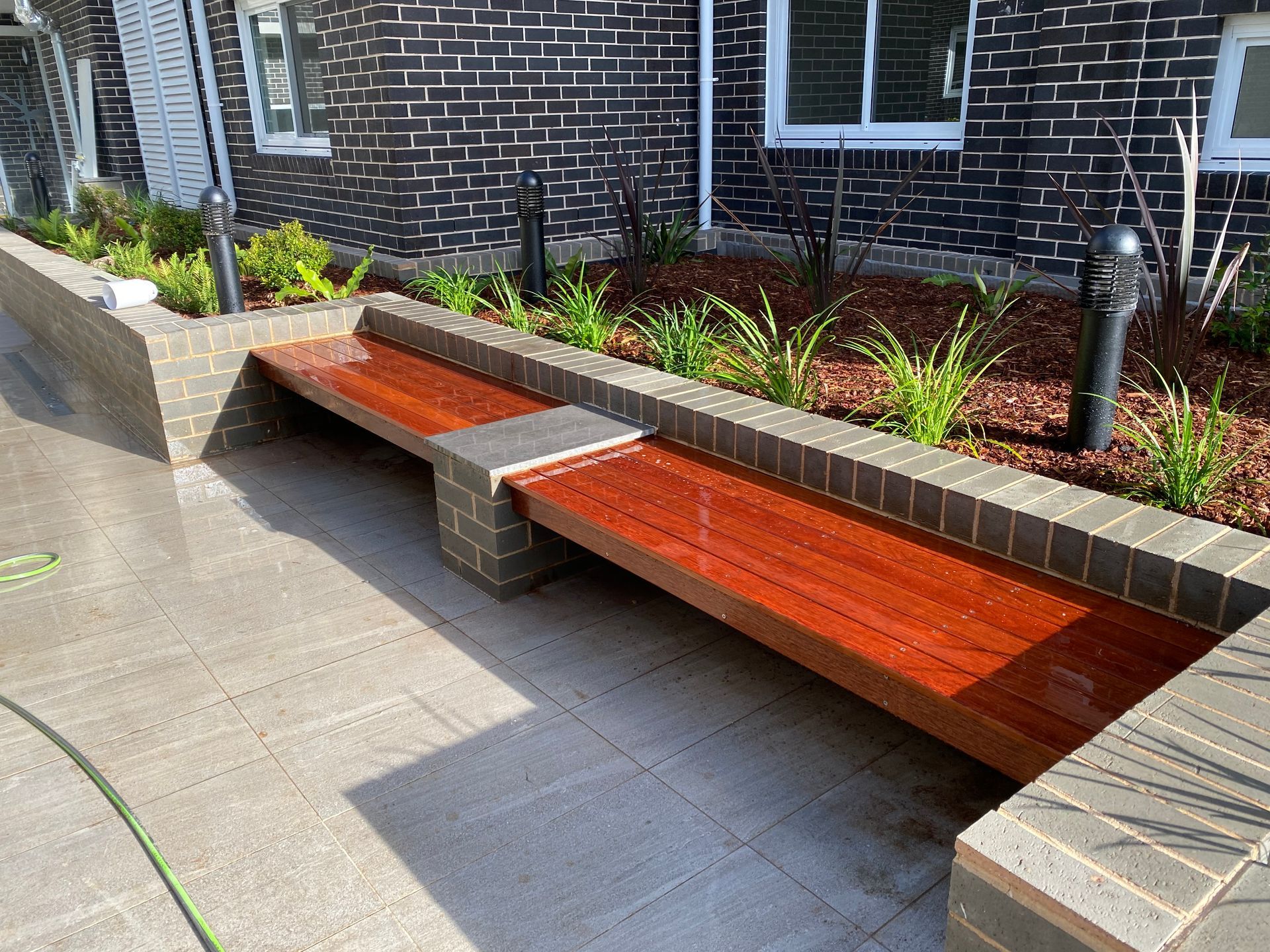 Wooden bench with brick accents, built into a garden bed with plants, against a brick building.