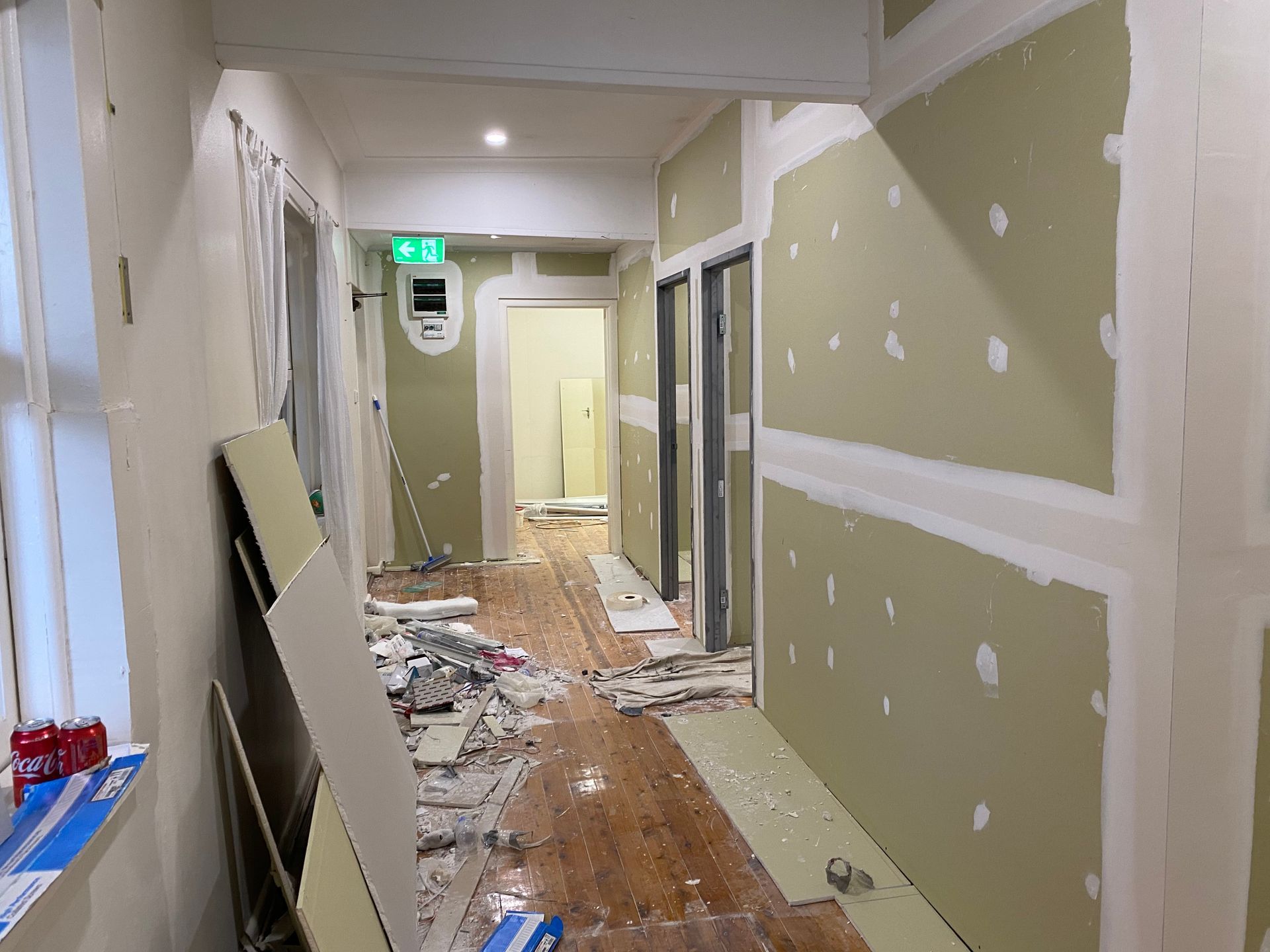 Hallway under renovation with exposed studs, drywall, and debris on wooden floor.