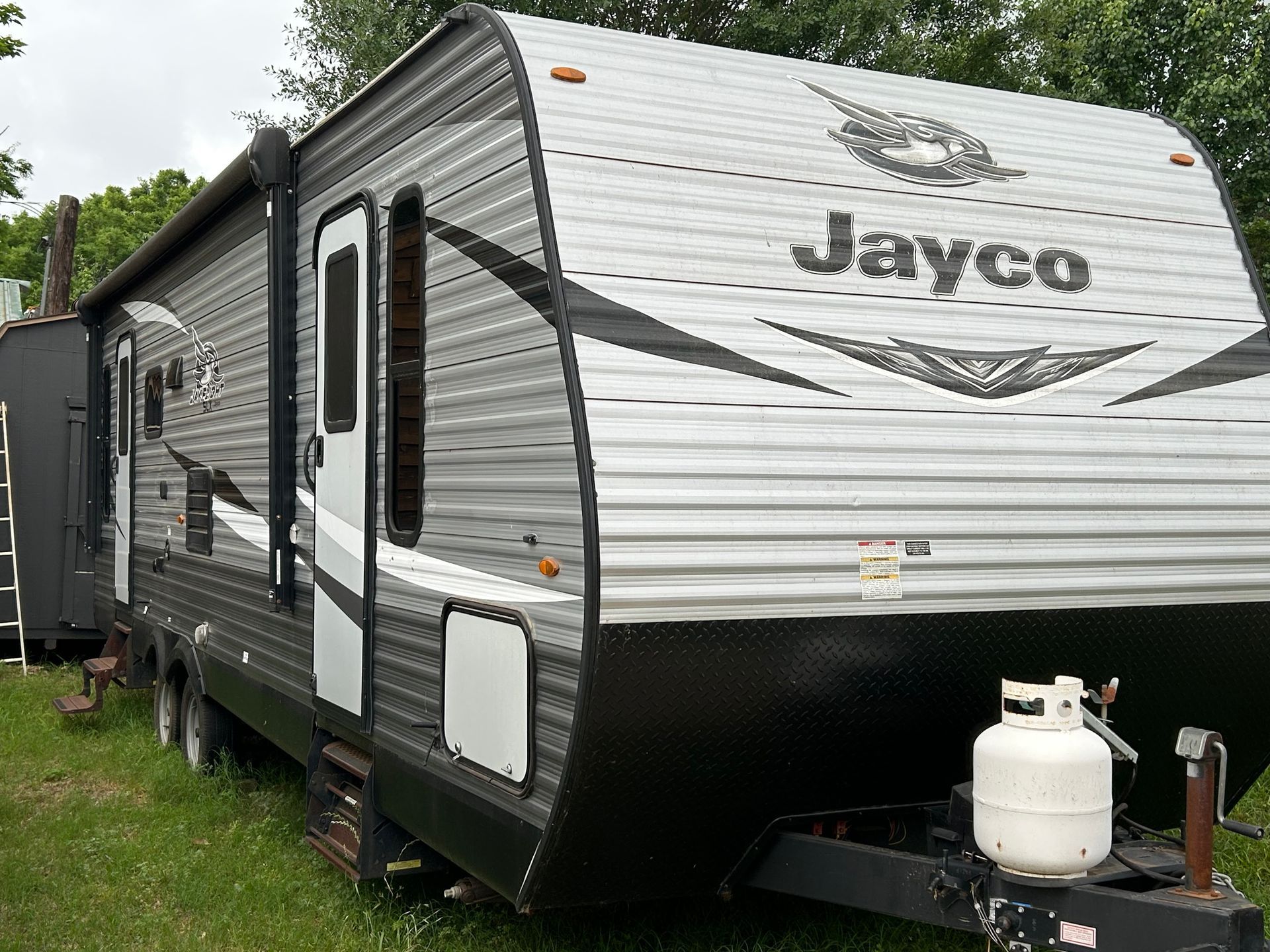 Gray and black Jayco travel trailer parked on grass, with propane tank in front.