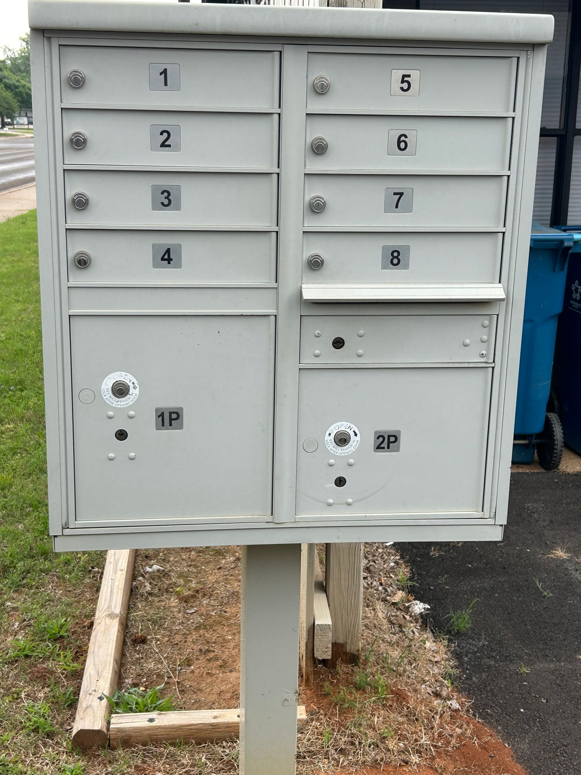 Mailboxes, gray metal, numbered 1-8, with two parcel compartments, mounted on a post outside.