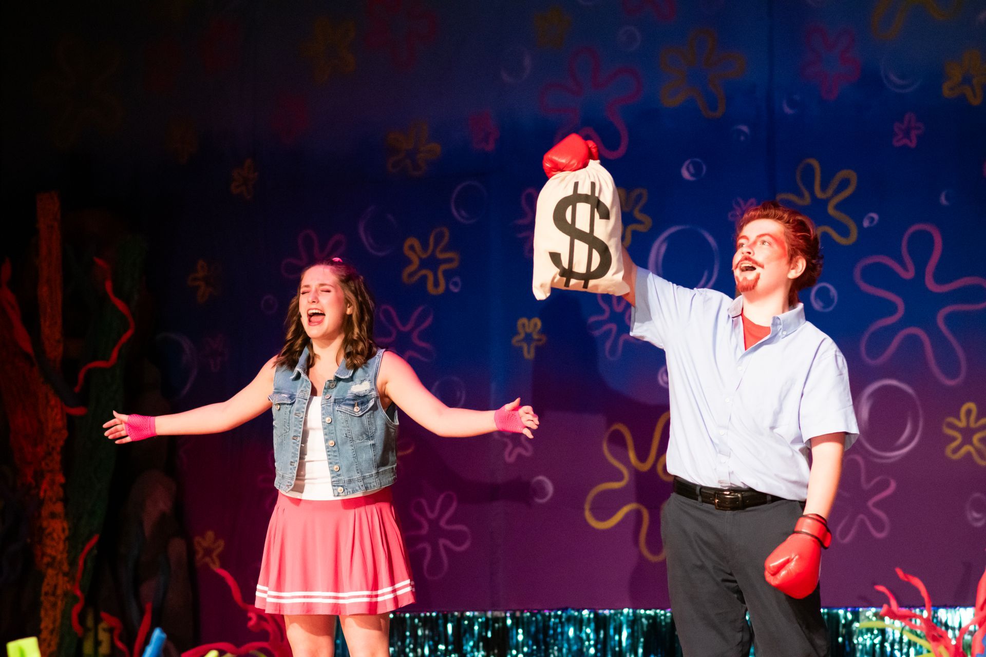 Two people on stage: woman in pink skirt, man holding money bag, both with boxing gloves.