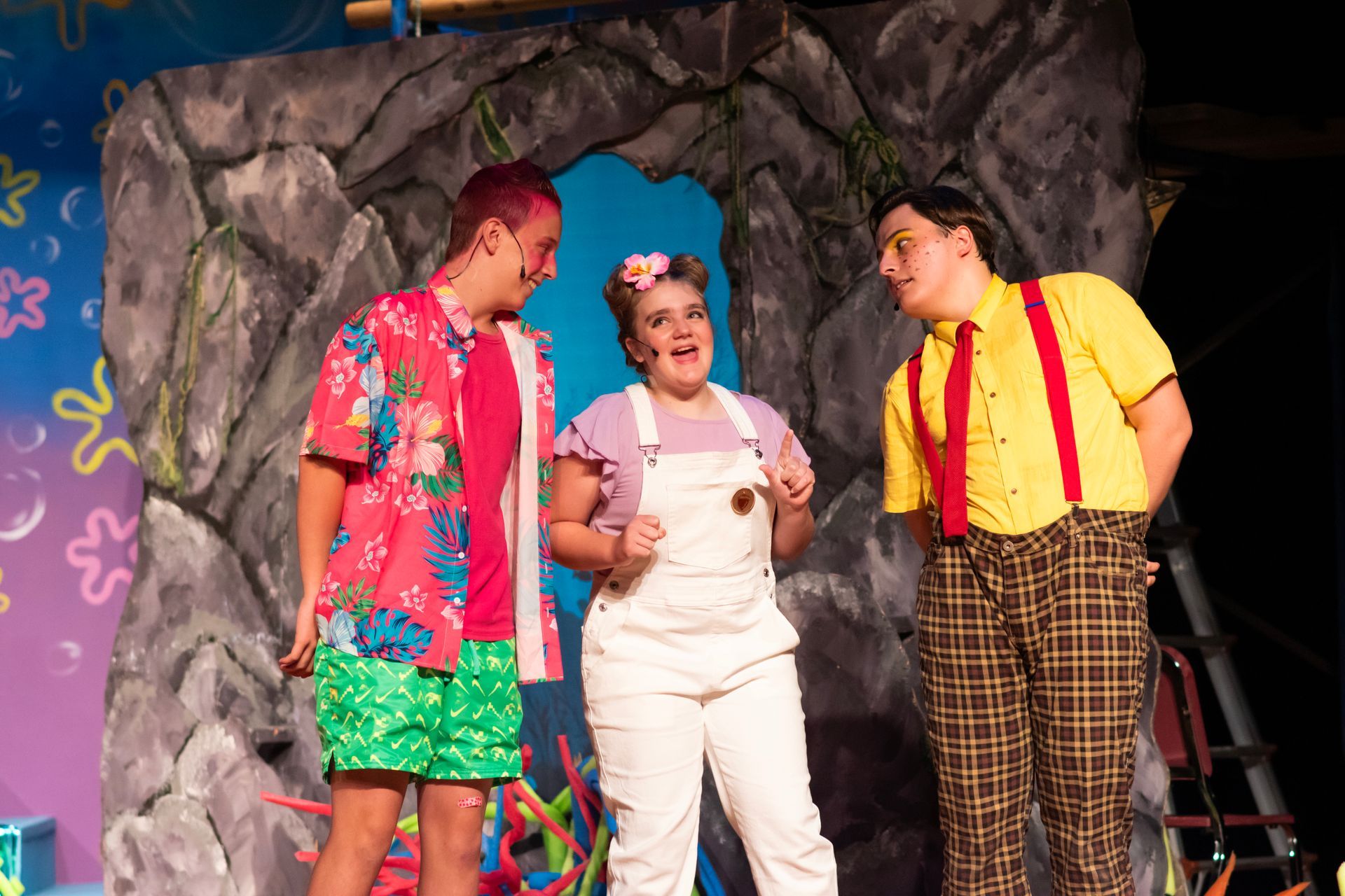 Three actors on a stage: one with pink hair in a Hawaiian shirt, one in overalls, and one in a yellow shirt and suspenders.